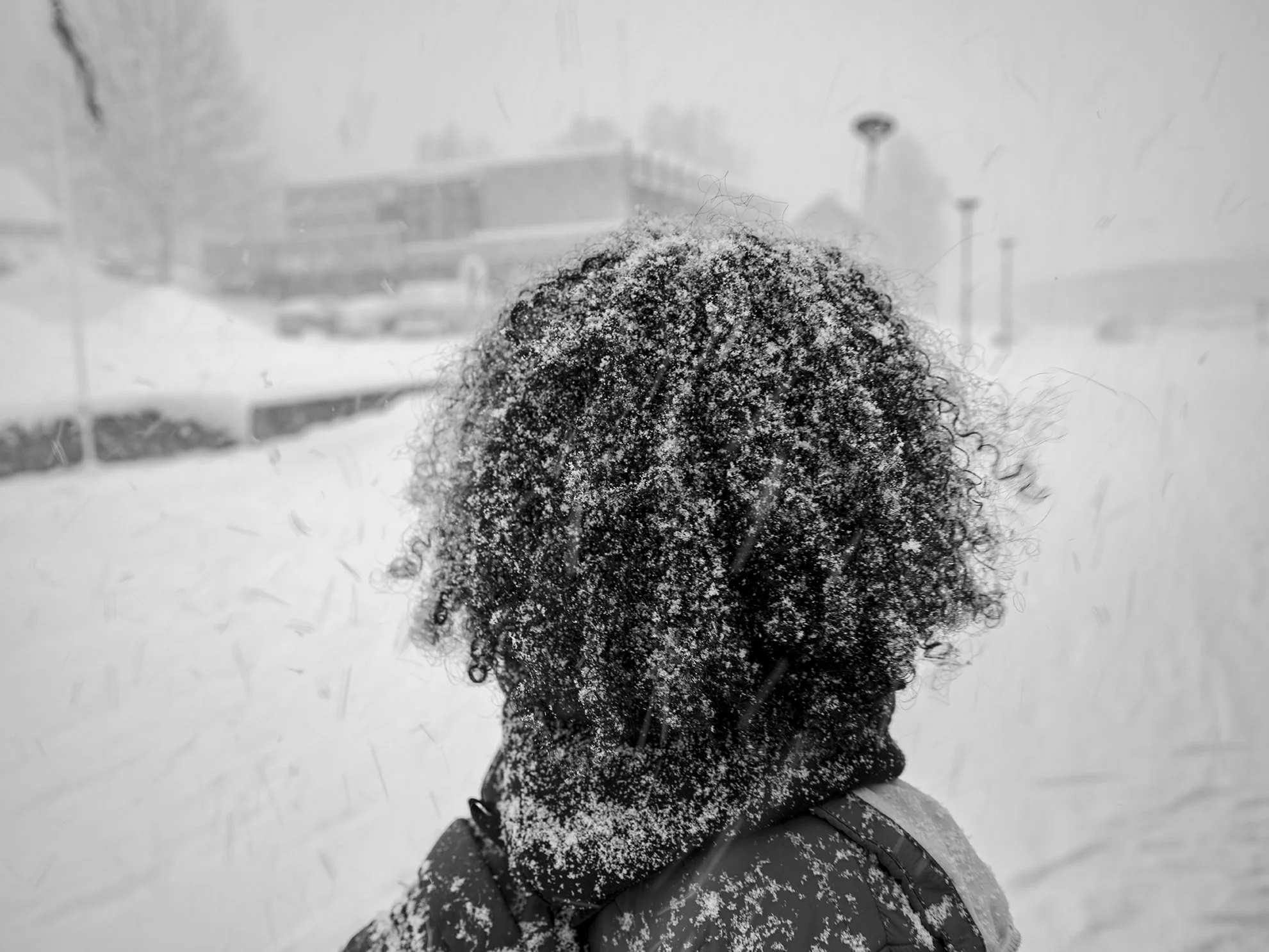 A person with curly hair covered in snow, standing outdoors during a snowstorm, with buildings and streetlights in the background.
