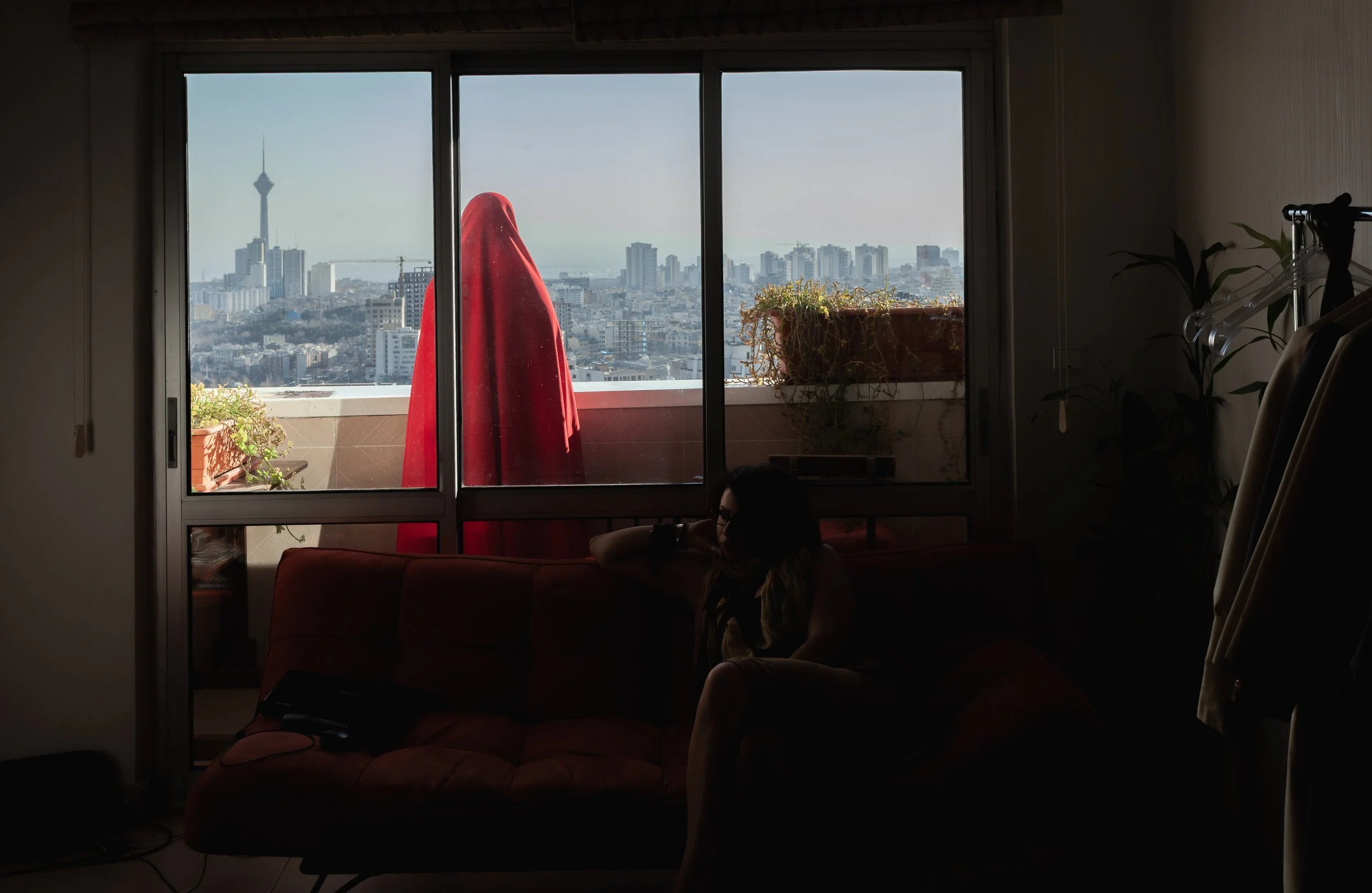 A woman sitting on a couch inside a dimly lit room, looking towards a large window with a cityscape view, including a tall tower in the distance. The outside balcony has a red curtain and potted plants.