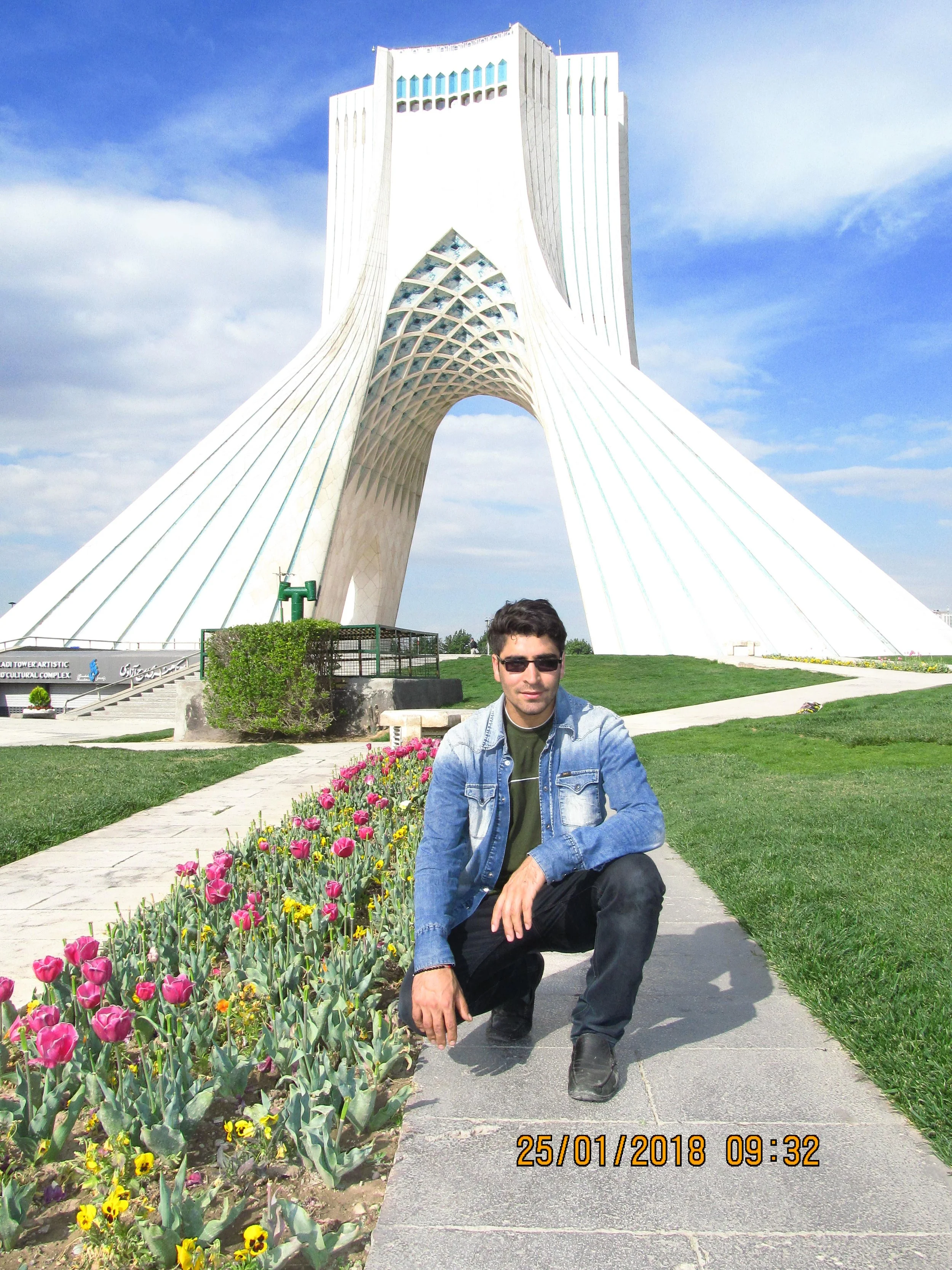 A man wearing sunglasses, a denim jacket, and black pants squatting on a sidewalk next to a flower bed with pink and yellow flowers, in front of a large, white, uniquely-shaped building with modern architecture against a partly cloudy sky.