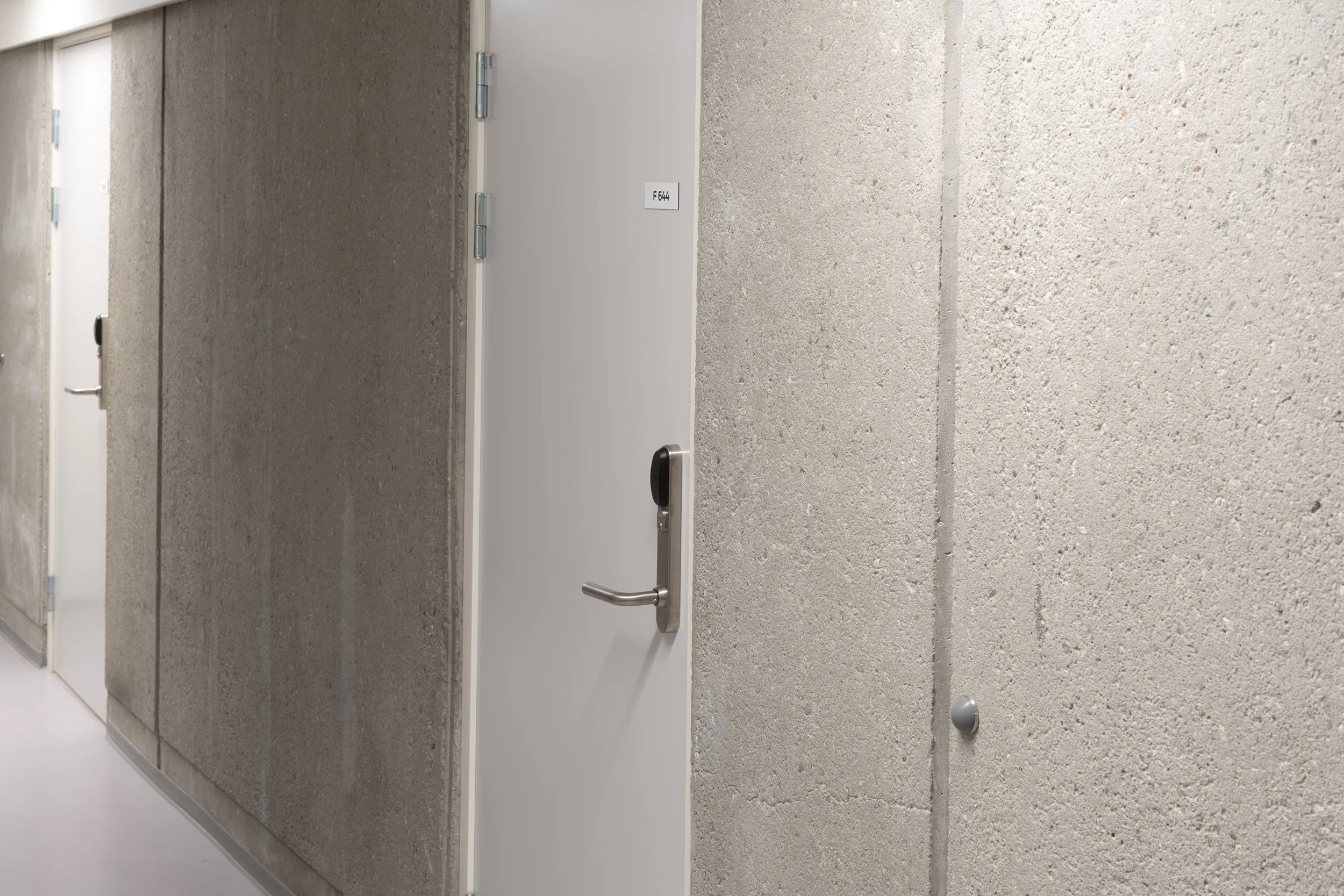 A corridor with textured concrete walls and a white door labeled F 644, featuring a silver handle and an electronic lock, inside a modern building.