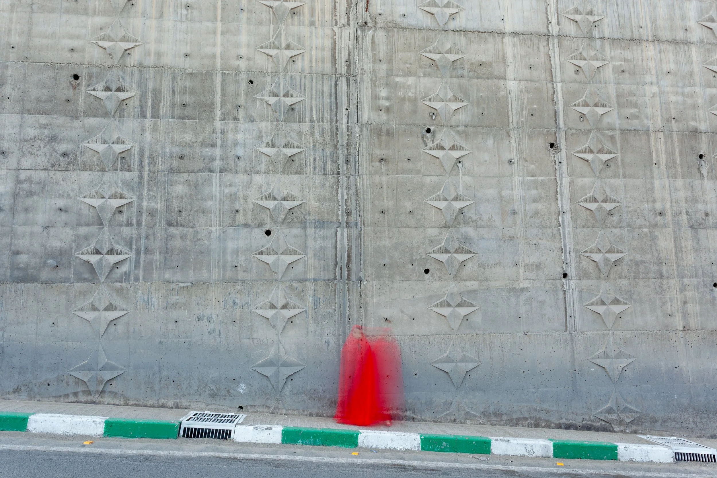Concrete wall with geometric diamond pattern and small holes, with a red spray-painted mark at the bottom near a curb painted in green and white stripes.