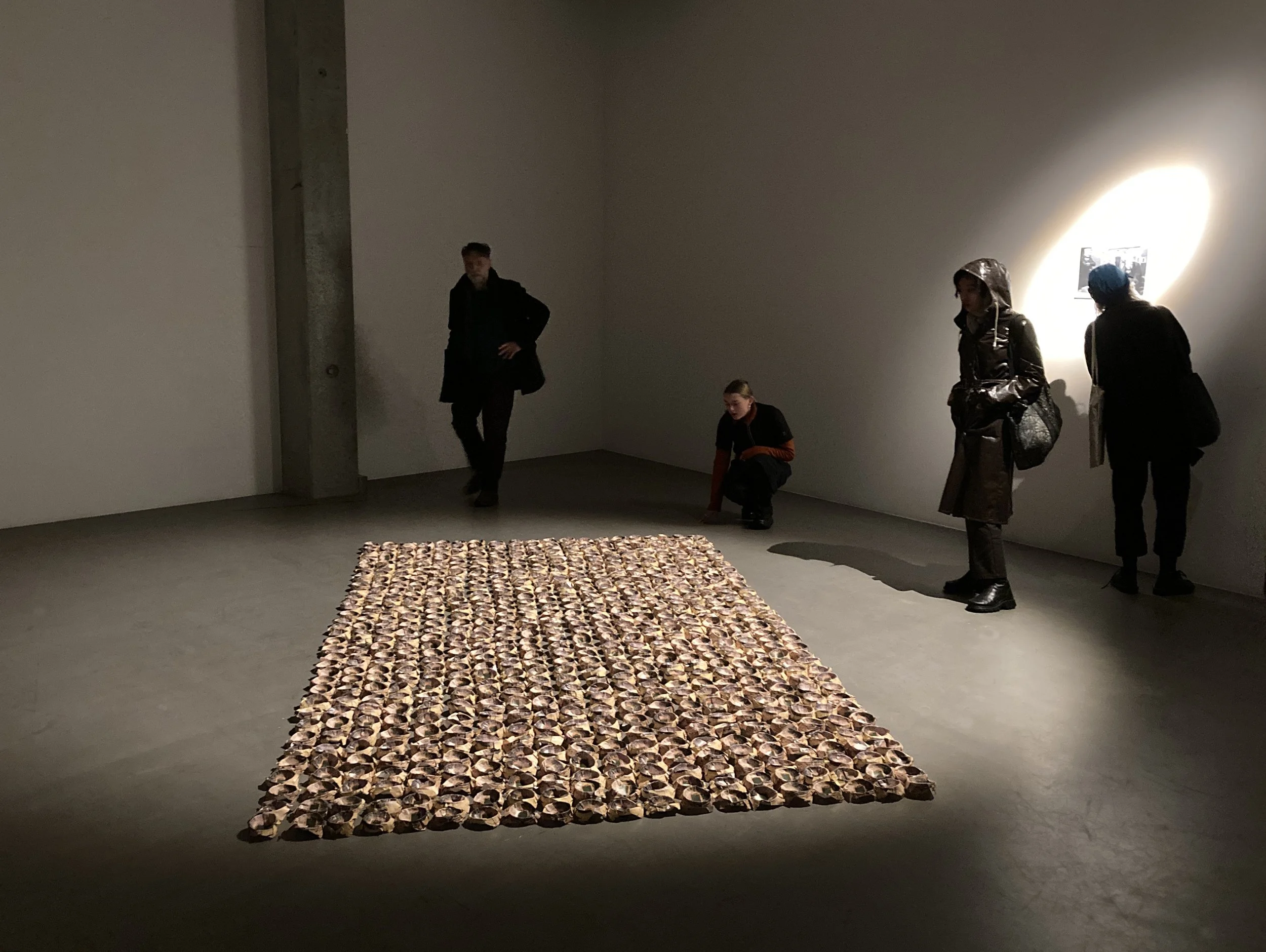 An art installation in a gallery with a large rectangular piece on the floor made of numerous small objects, and four visitors observing the artwork.