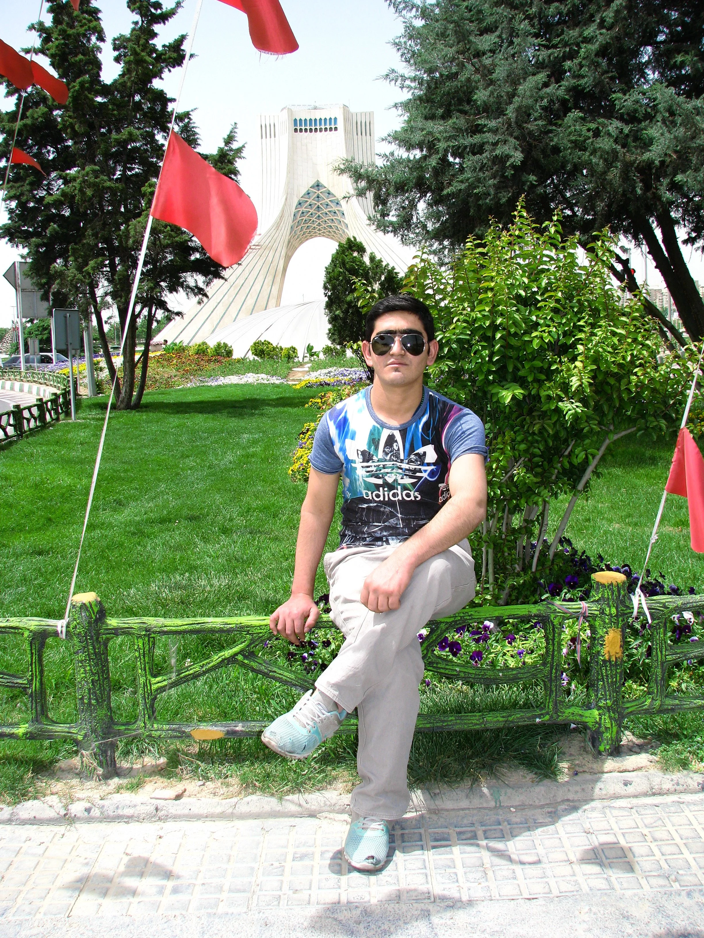 A young man wearing sunglasses, a graphic t-shirt, light-colored pants, and sneakers sitting on a decorative green metal bench in a park with the Azadi Tower in the background.