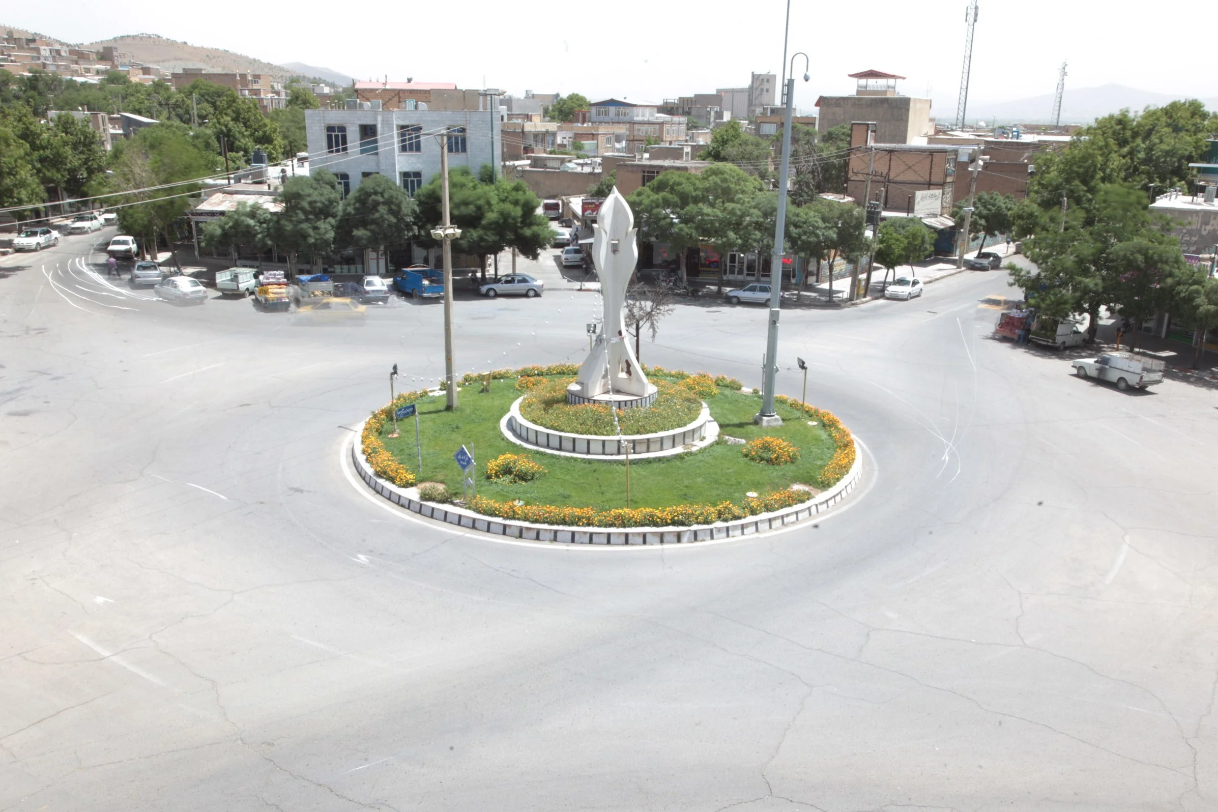 A roundabout with a modern sculpture at the center, surrounded by a flower bed and several trees, in a small town or city with buildings, parked cars, and power lines in the background.