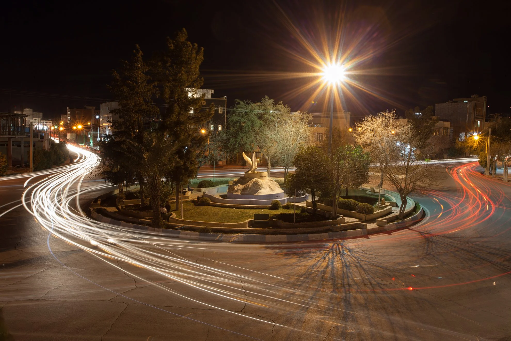 Long exposure photo of a city roundabout at night with light trails from moving vehicles, a small fountain with a sculpture of a bird, trees, and buildings in the background.
