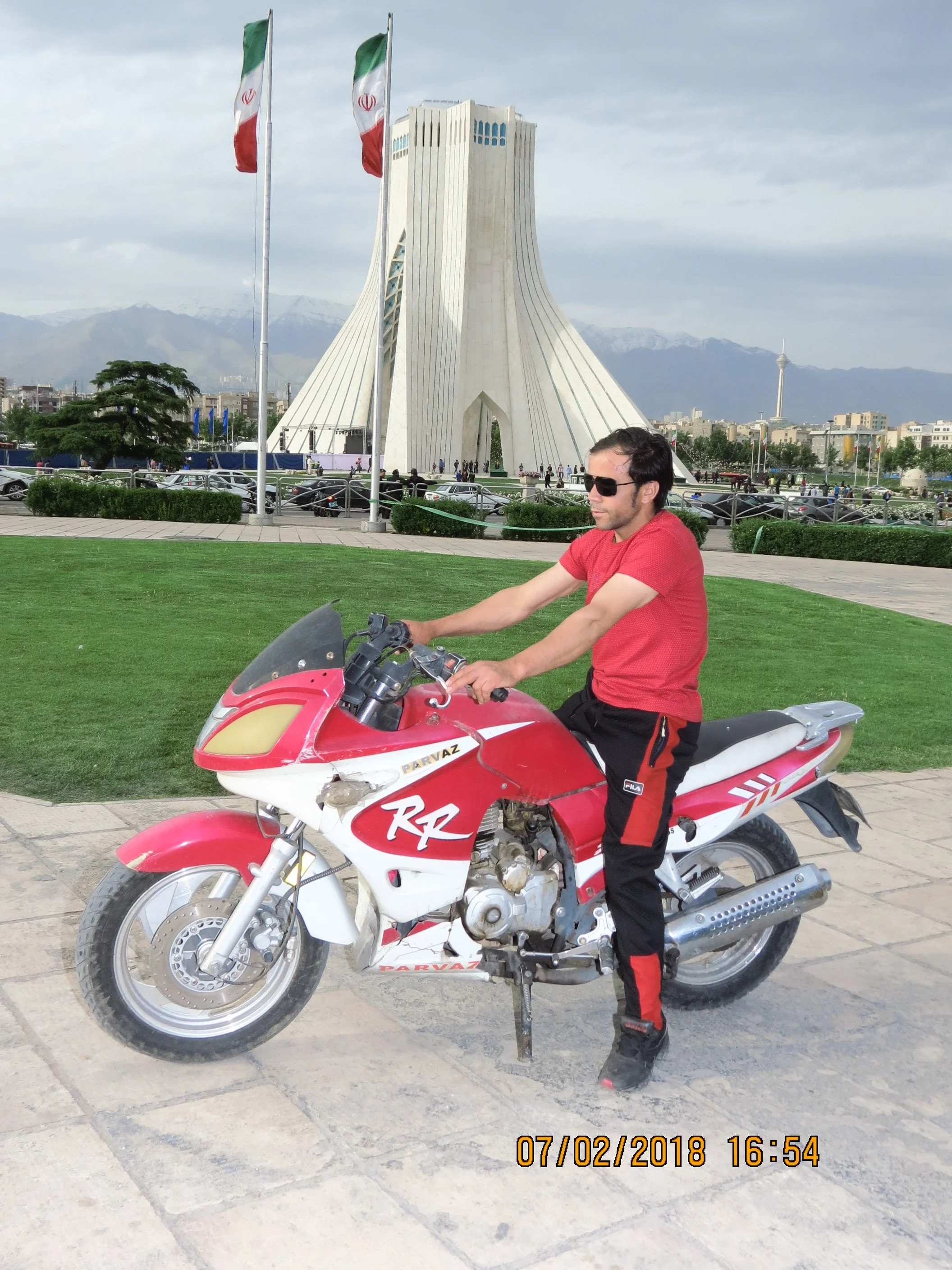 A man in a red T-shirt and black and red pants sitting on a red and white motorcycle in front of a monument with flags in the background.