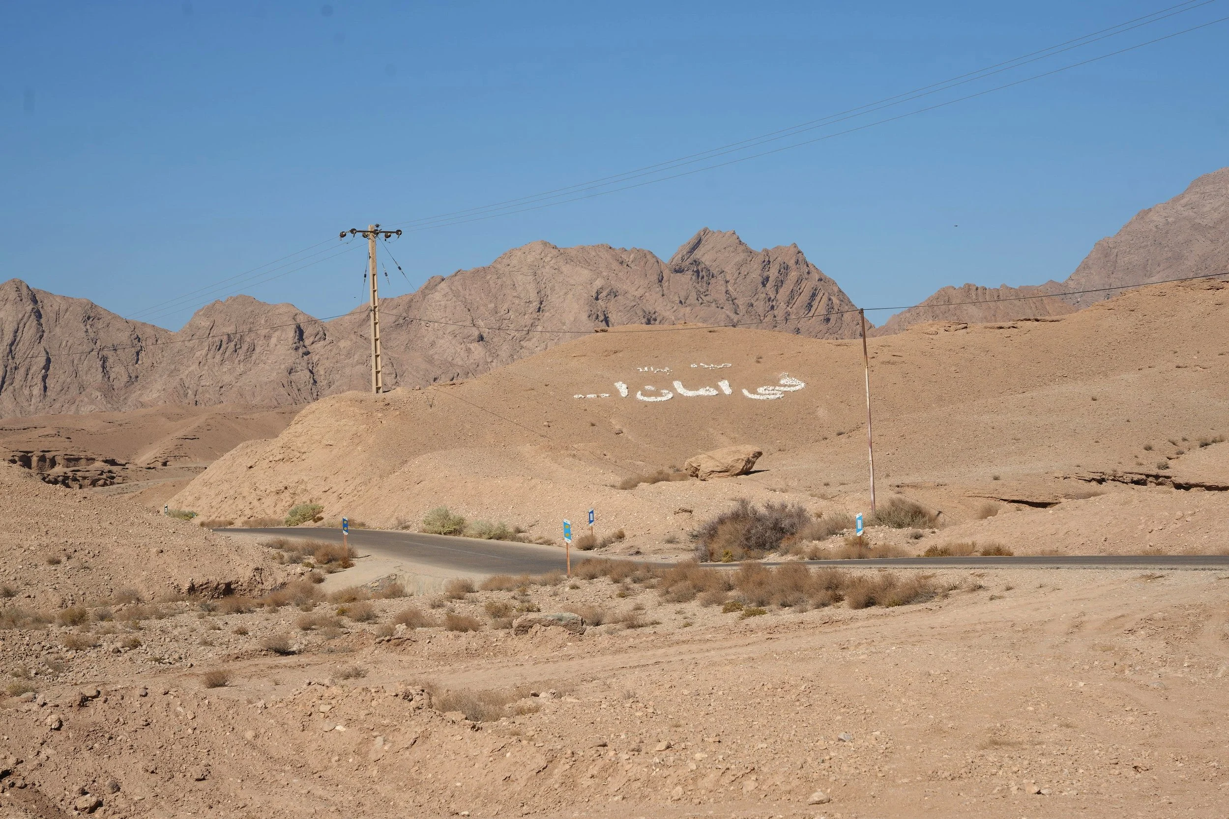 Desert landscape with mountains in the background, a winding road, and rock-formed hills. There is Arabic writing on one of the hills and utility poles along the road.