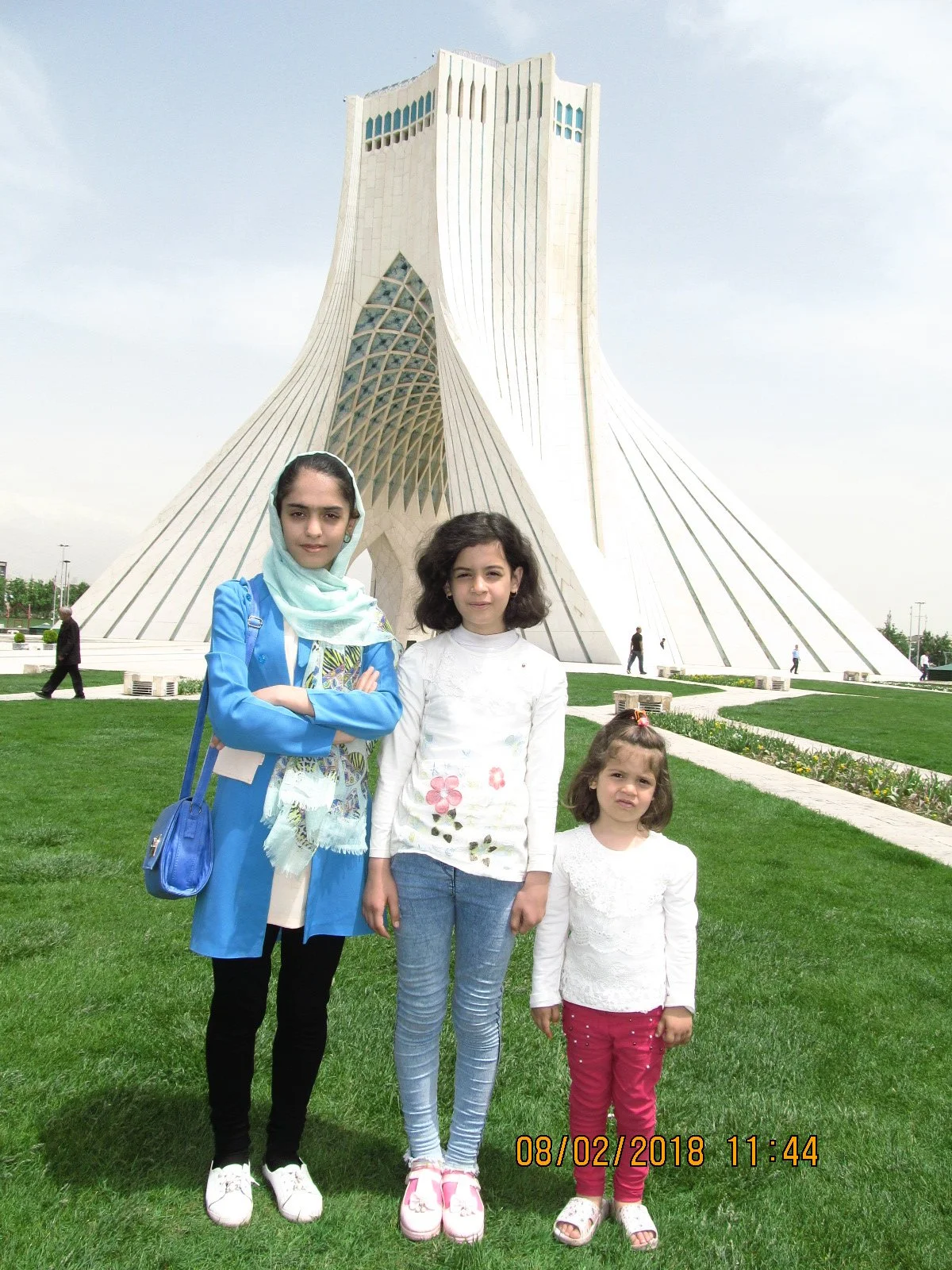 Three young girls standing in front of the Azadi Tower in Tehran, Iran, on a grassy area with a modern architectural building in the background.