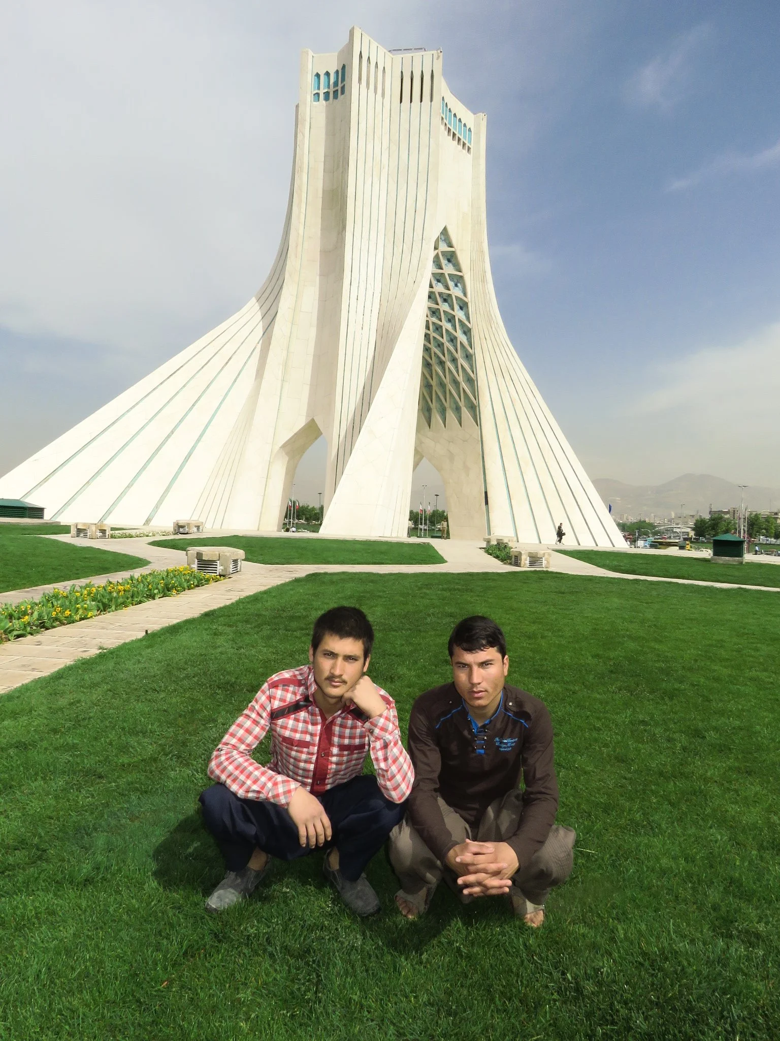 Two young men squatting on a well-maintained grassy area in front of the Azadi Tower in Tehran, Iran. The tower is a tall, white, architecturally unique monument with a modern design against a partly cloudy sky.