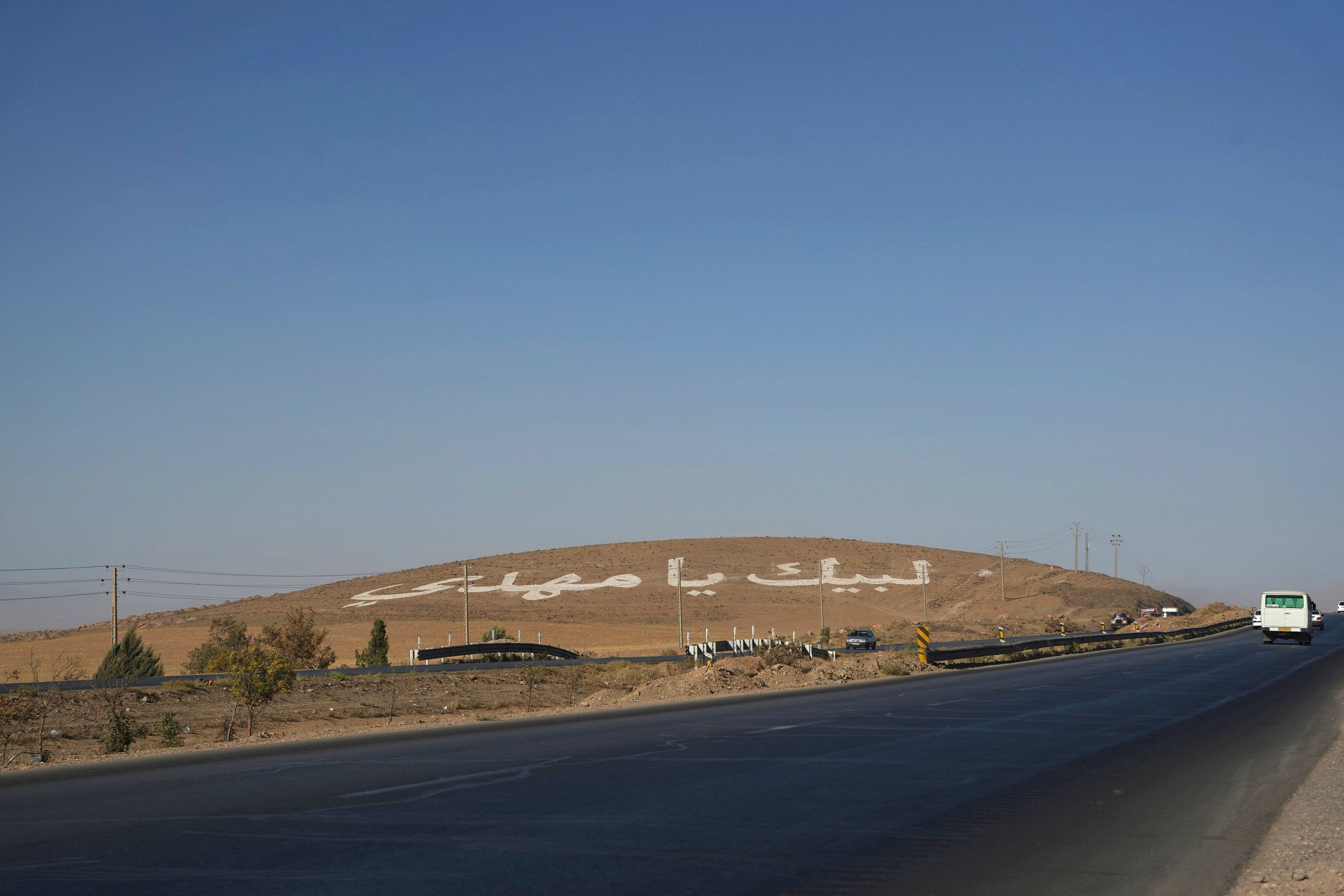A hillside with white letters spelling a message in Persian, with a road and cars in the foreground.