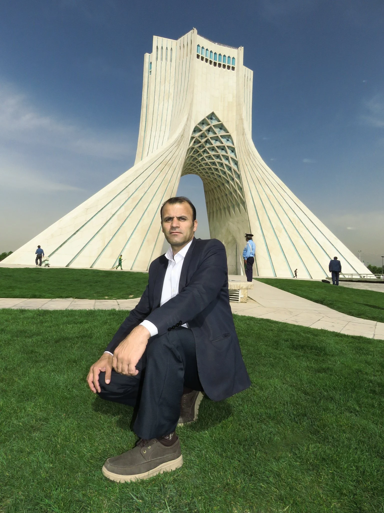 A man dressed in a suit squatting on grass in front of a modern architectural building with a distinctive arch and geometric design, under a cloudy sky.