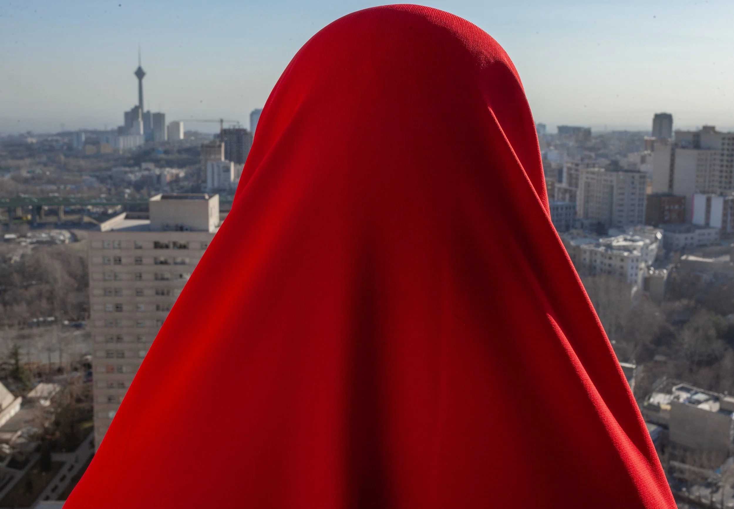 A person wearing a red hooded garment standing on a balcony overlooking a cityscape with tall buildings and a tower in the distance