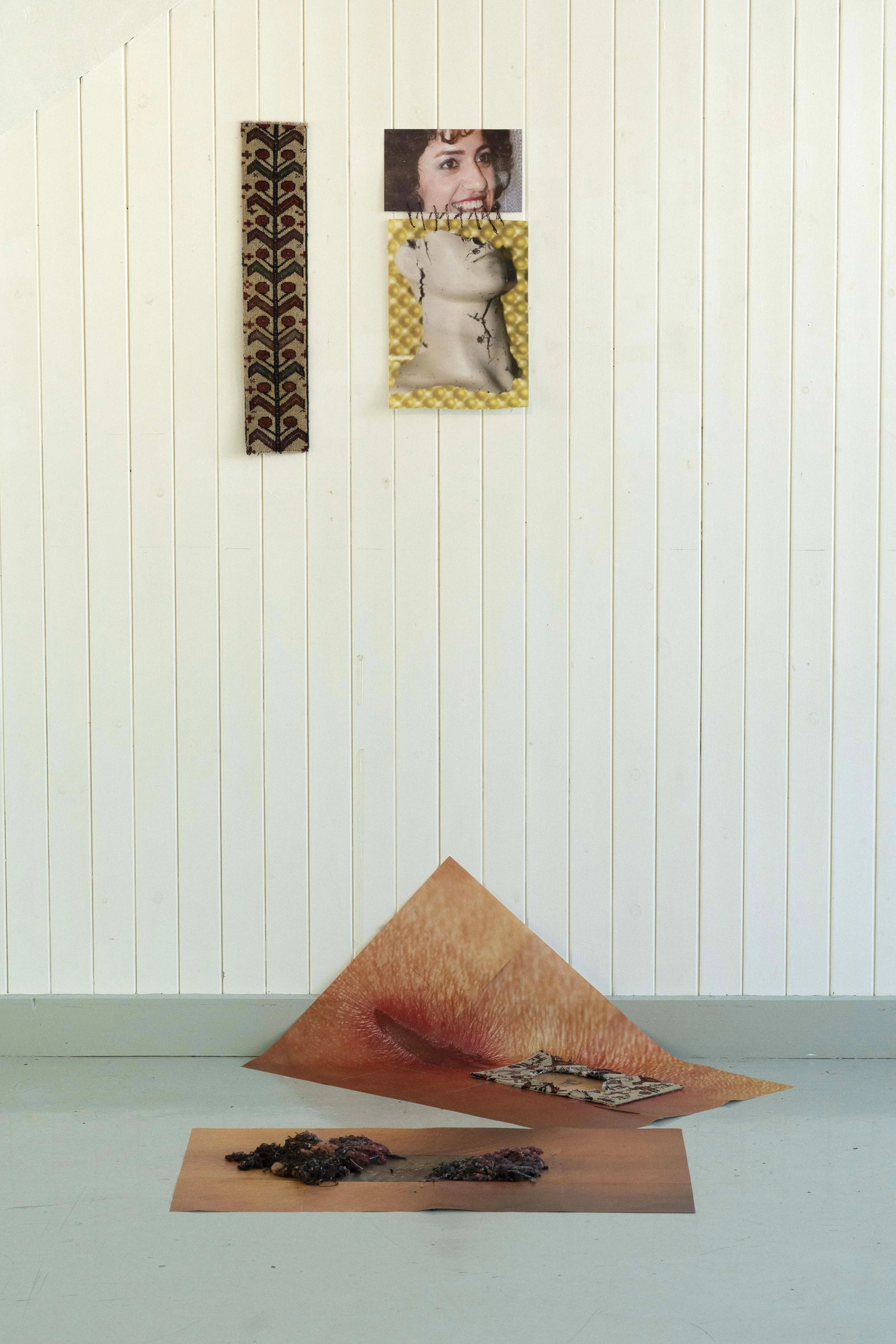 An art installation featuring collaged images and textured materials. A picture of a smiling woman’s face is mounted on the wall. Below it, a duct tape-covered face with cracked paint is visible. On the floor, there are large folded sheets of paper with photographic images, including a close-up of lips and a tiny image of a landscape or cityscape, alongside dark, textured material resembling burnt or charred debris.