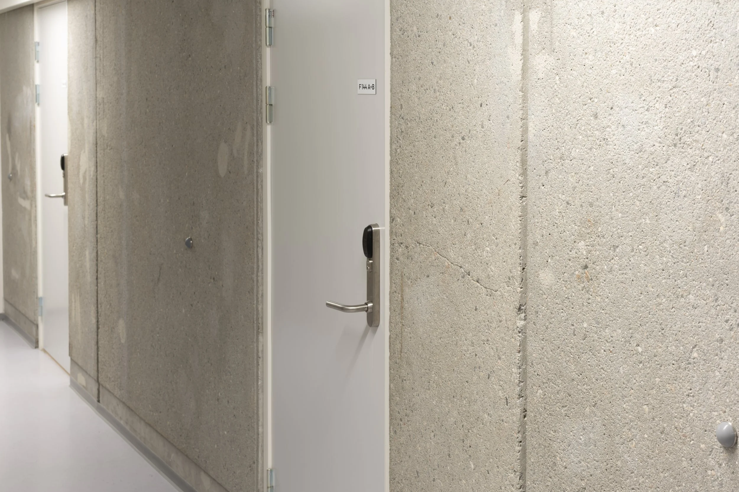 Corridor with concrete walls and multiple white doors, one of which has a handle and a small sign.