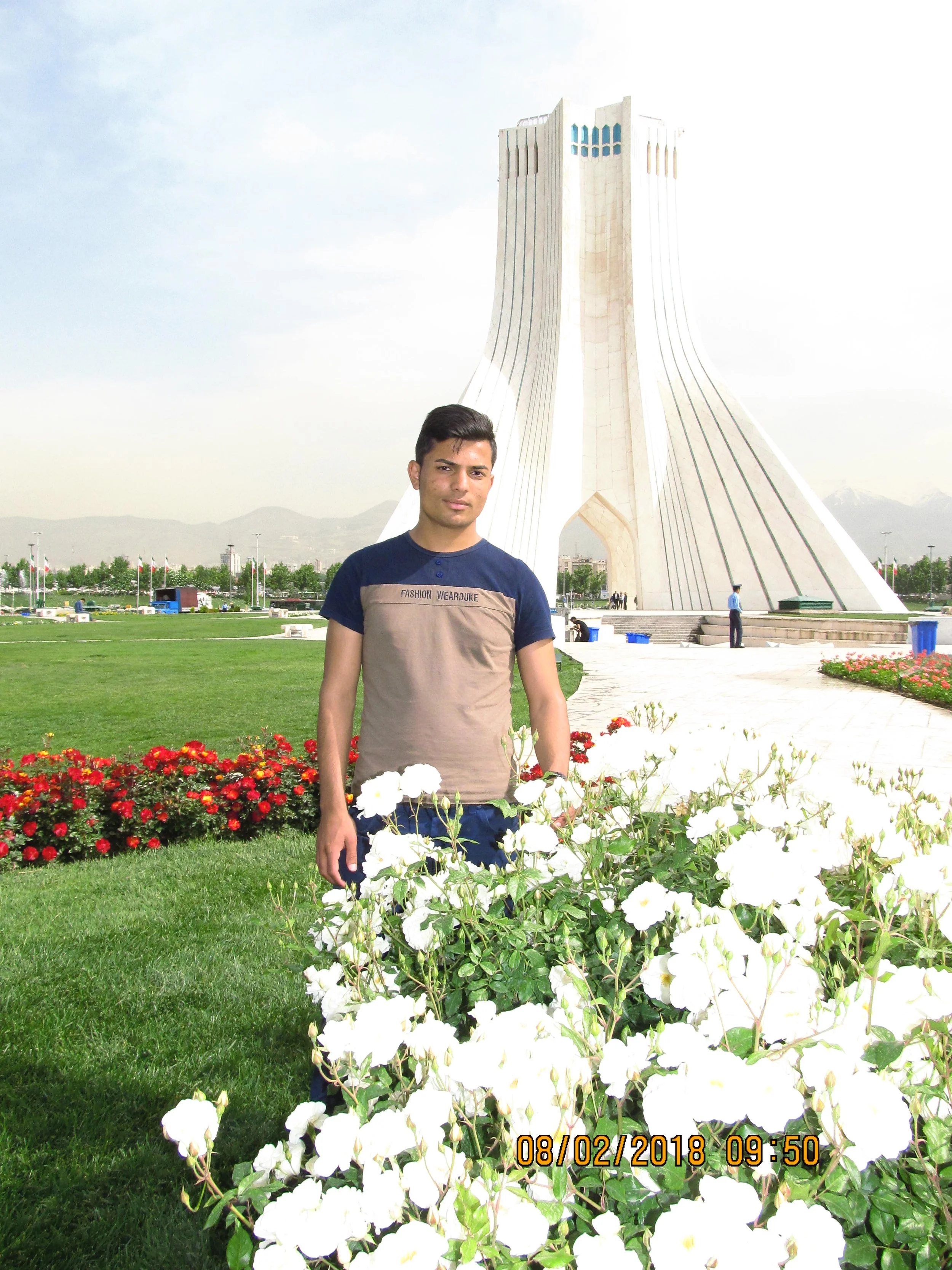A young man standing in a garden with white flowers, with the Azadi Tower in Tehran, Iran in the background.