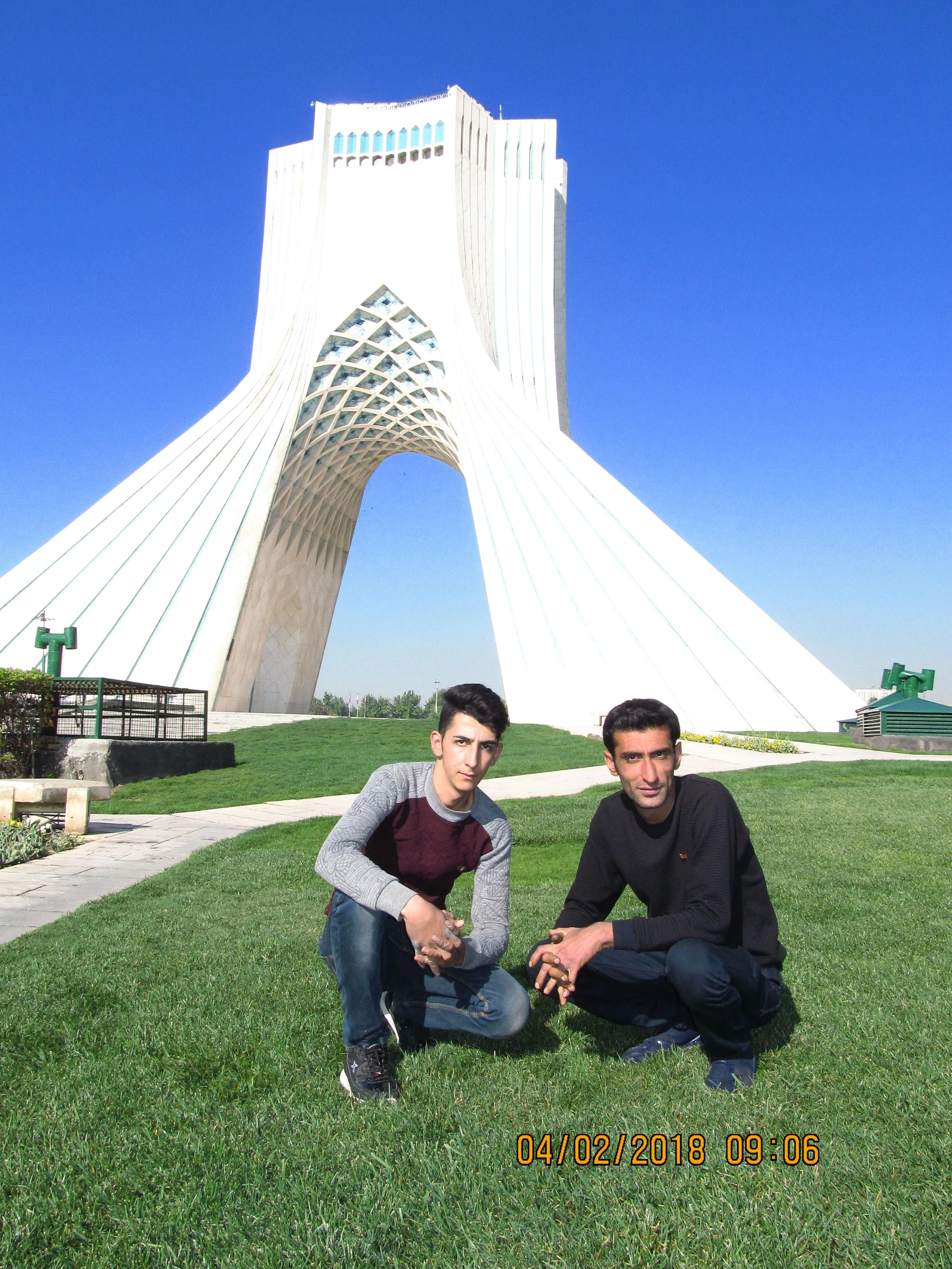 Two young men crouching on the grass in front of Azadi Tower in Tehran, Iran, under clear blue sky.