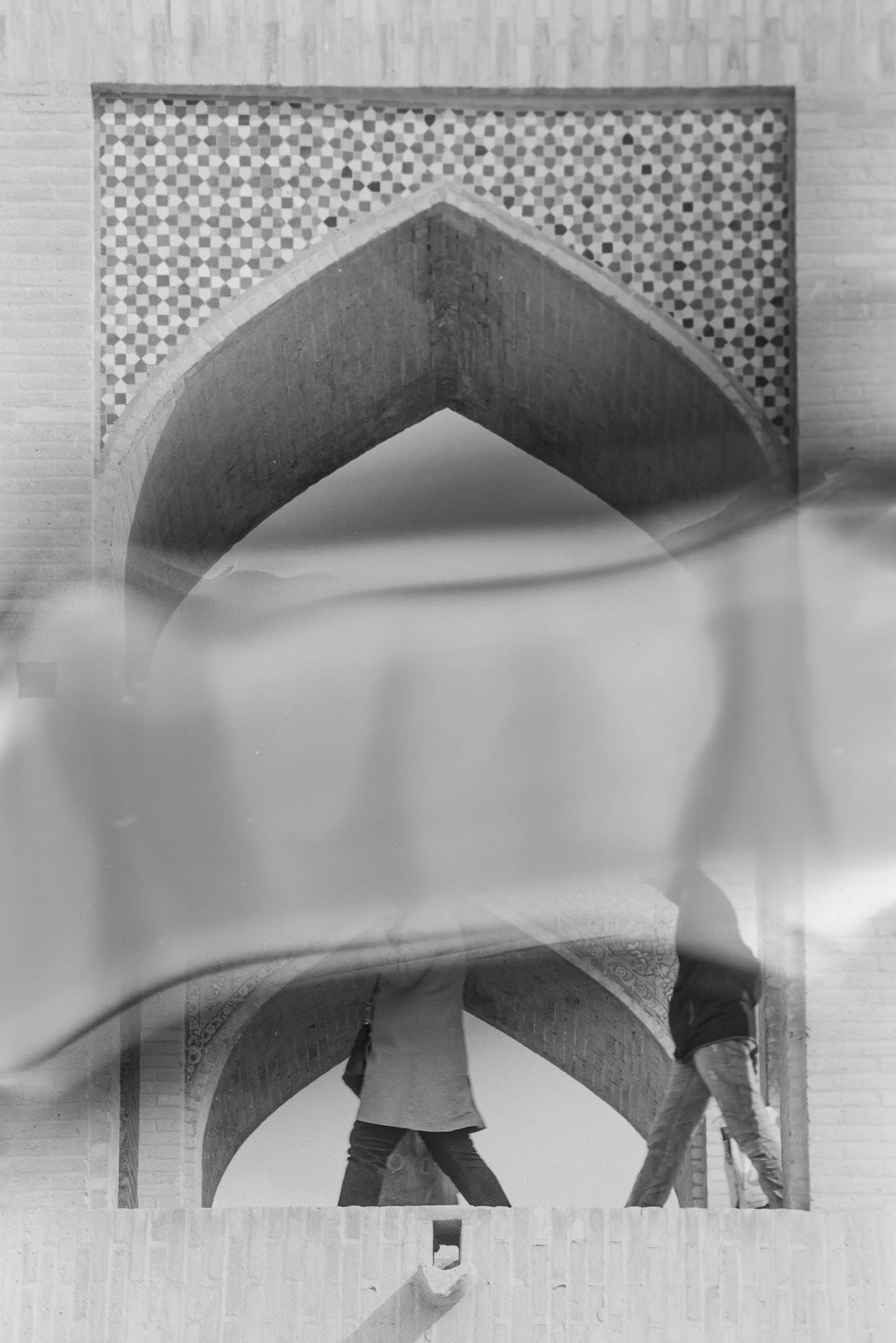 Black and white photo showing a reflected image of two people walking on a bridge with arched openings, with the main building above containing patterned tiles and brickwork.