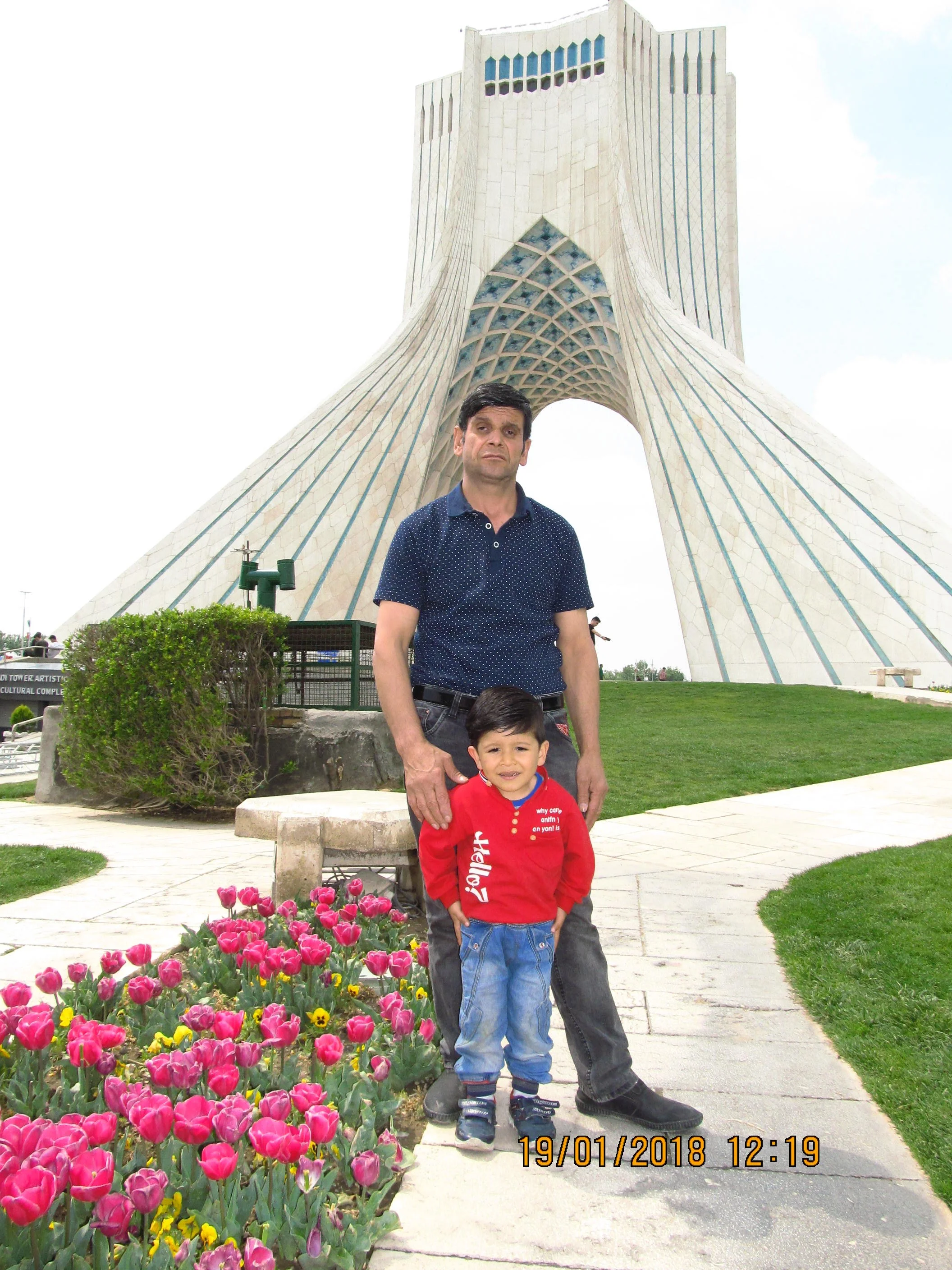A man and a young boy stand in front of Azadi Tower in Iran during daytime, with pink tulips and green grass around them.