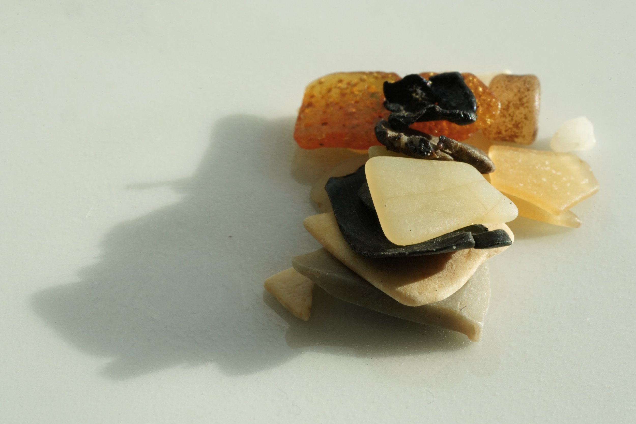 A collection of assorted sea stones and shells arranged on a white surface, with shadows cast to the left from sunlight.