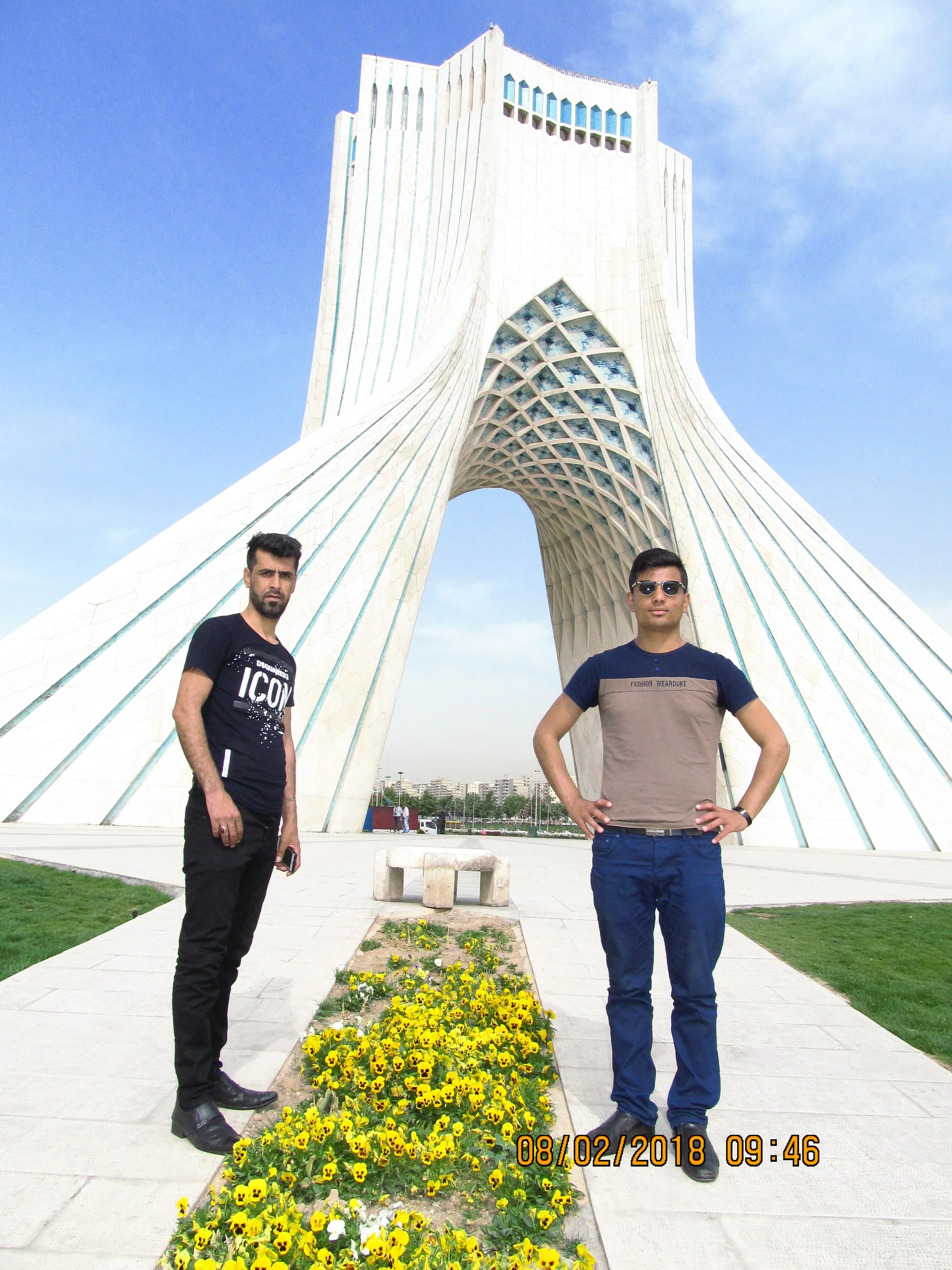 Two young men standing in front of the Azadi Tower, a large white monument with intricate lattice details and an archway, under a blue sky with some clouds. Flower bed with yellow flowers is in the foreground.