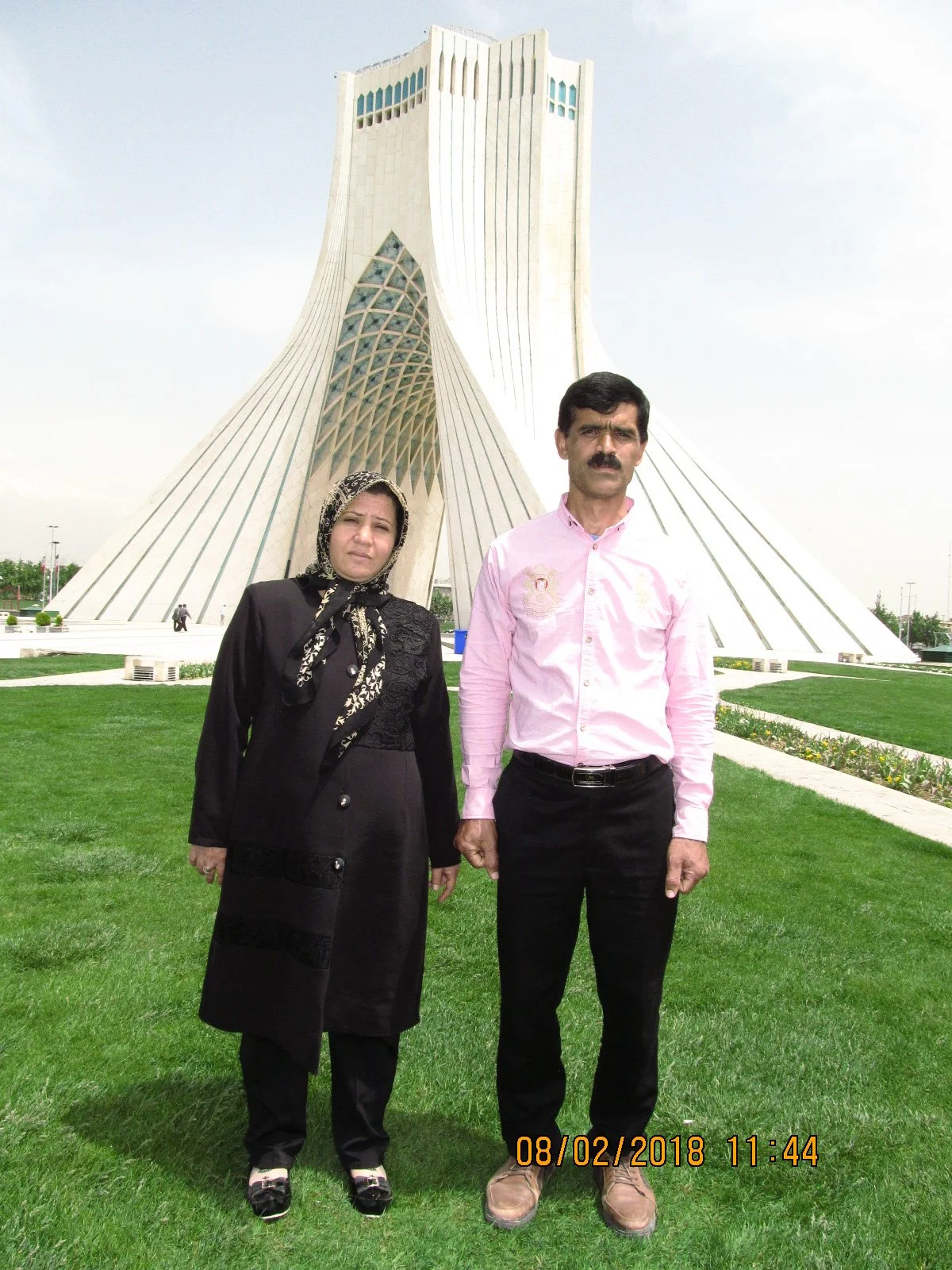 A man and a woman standing on a grassy area in front of a large modern building with a unique architectural design. The woman is dressed in black, wearing a headscarf, and the man is wearing a pink shirt and black pants.
