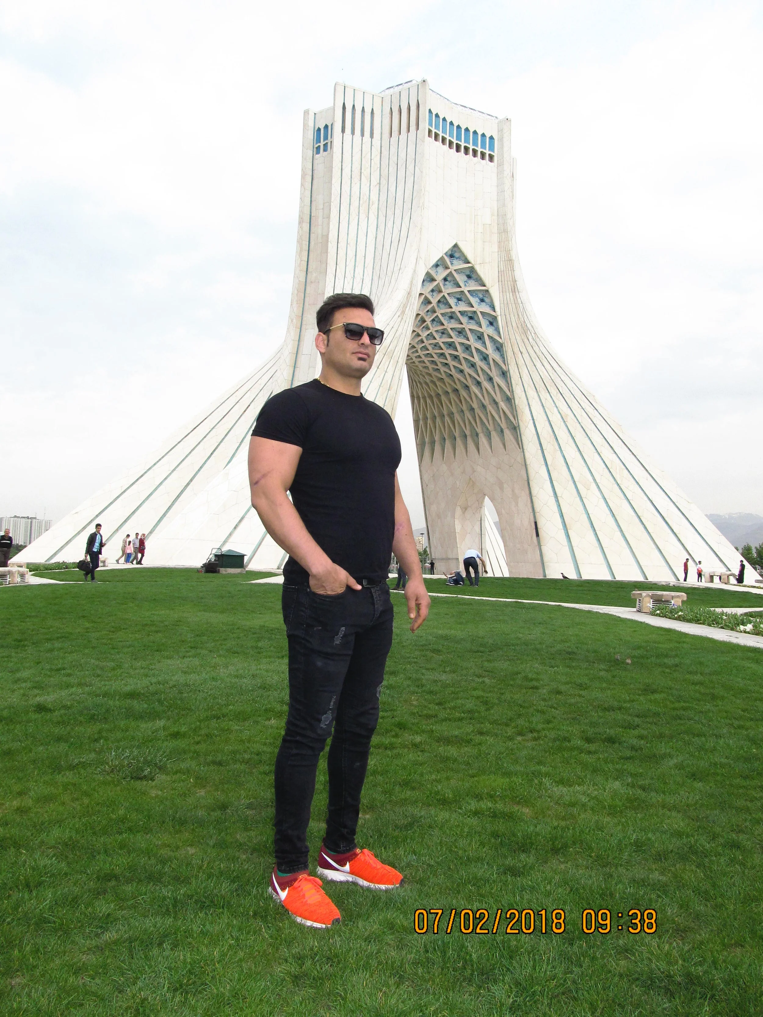 A man with sunglasses wearing a black t-shirt and ripped black jeans standing on green grass in front of the Azadi Tower in Tehran, Iran.