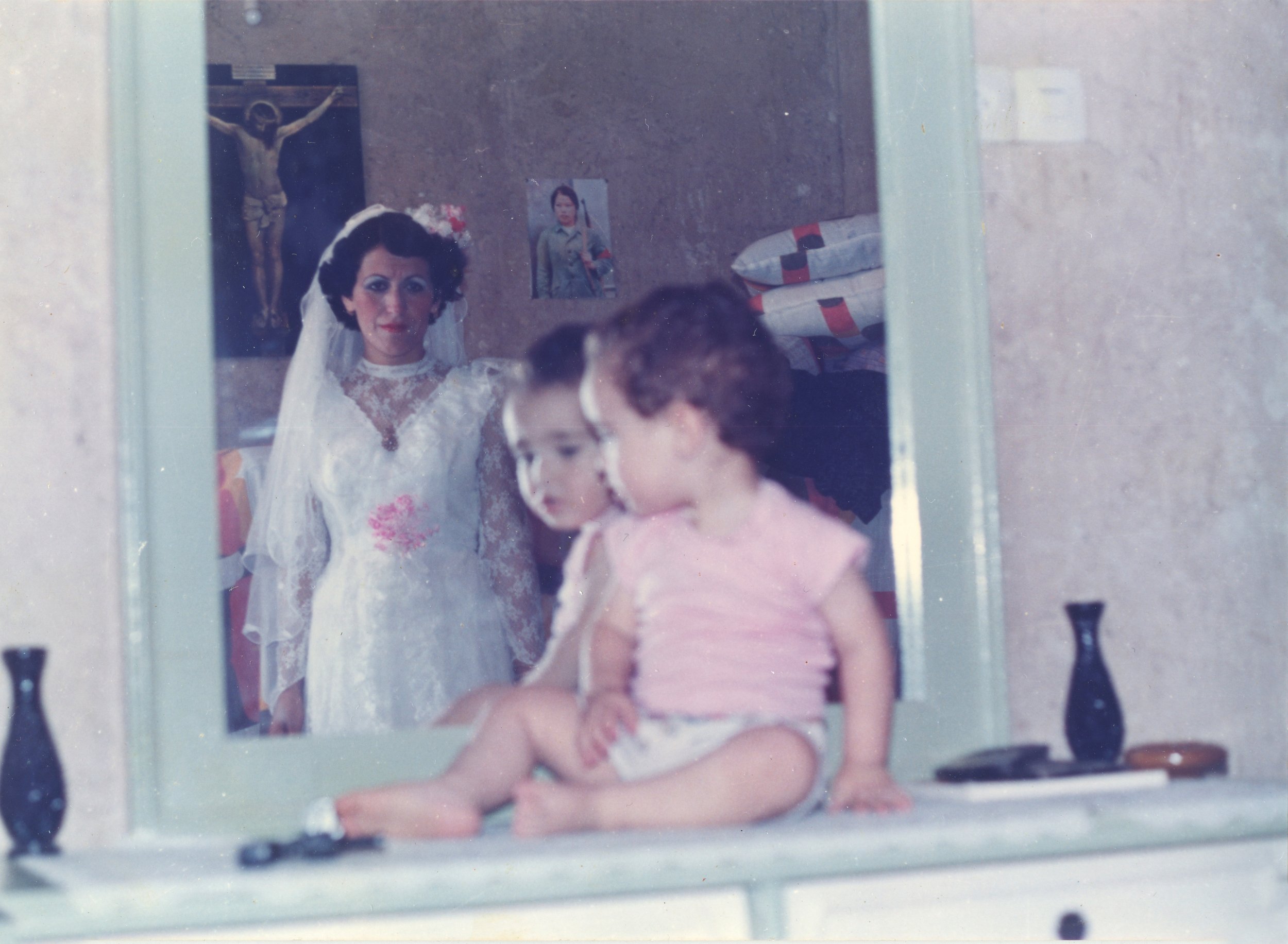 A bride in a white wedding dress and veil looking into a mirror, with two children sitting on a dresser in front of her, one wearing a pink shirt and the other with a white shirt and dark hair. Religious and portrait posters on the wall behind.