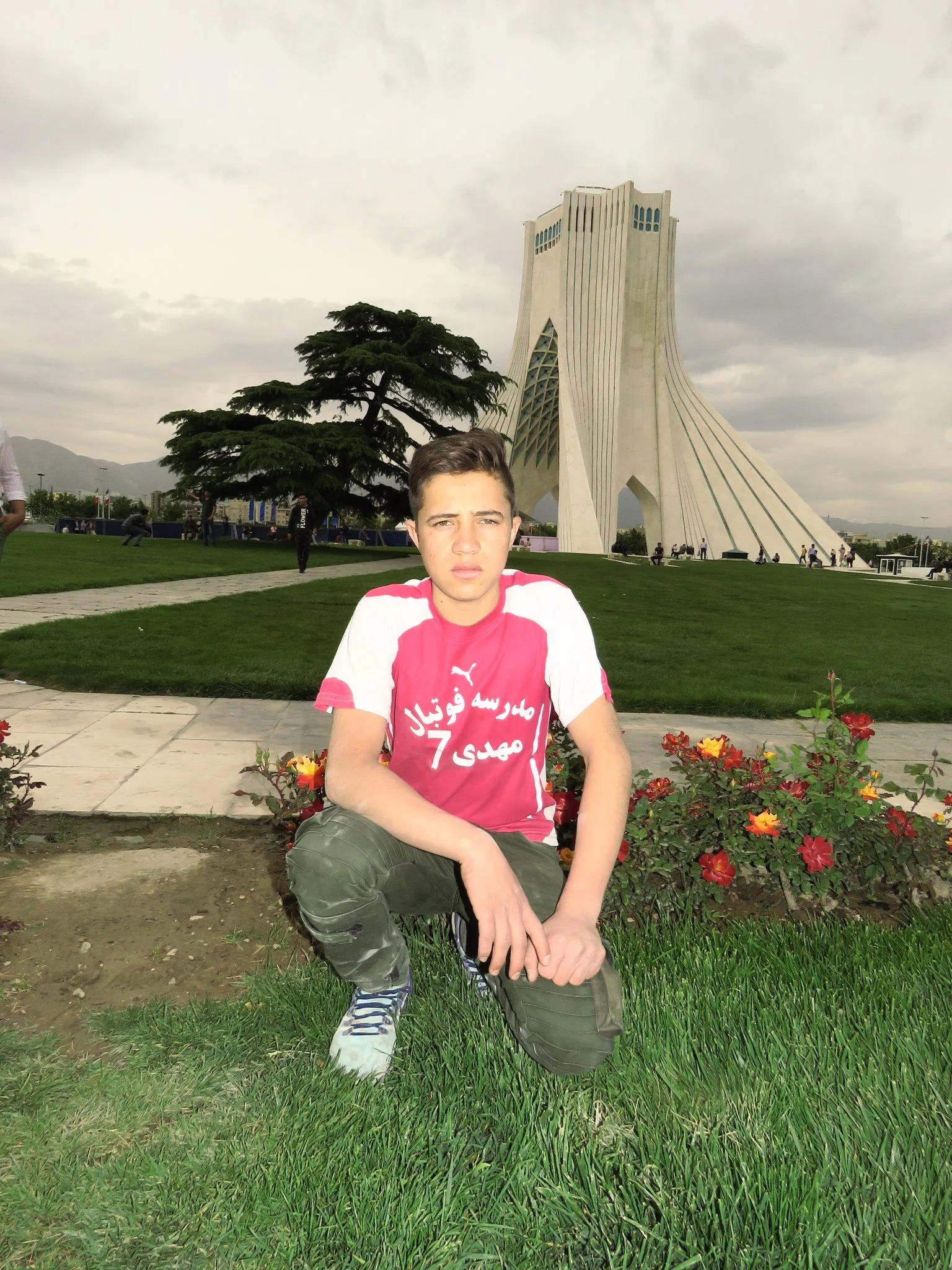 A young boy in a red and white sports jersey crouches in a garden with flowers, with a modern architectural building, trees, and people in the background on a cloudy day.