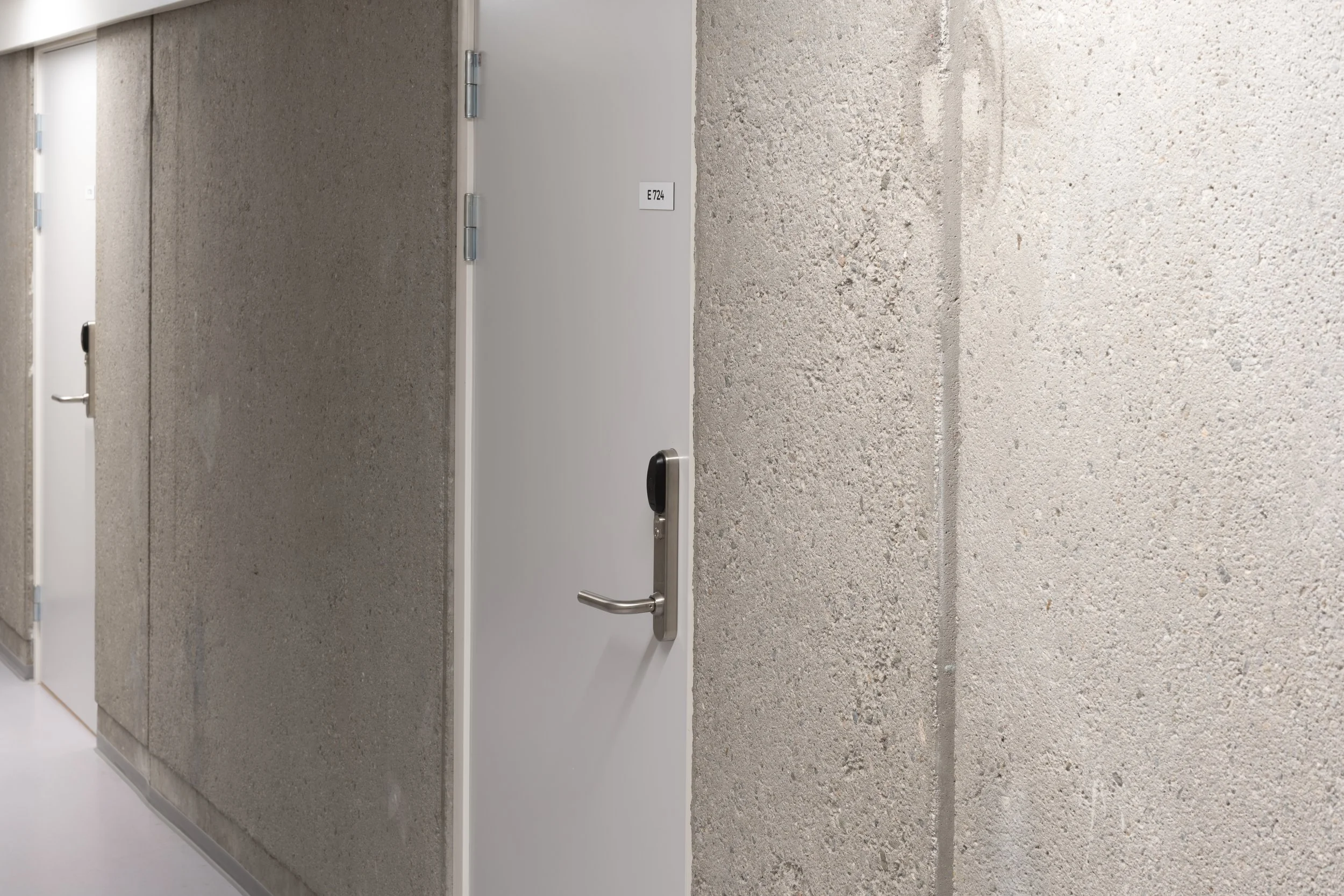 A hallway with textured concrete walls and white doors, one door has a black handle and a room number sign E724.