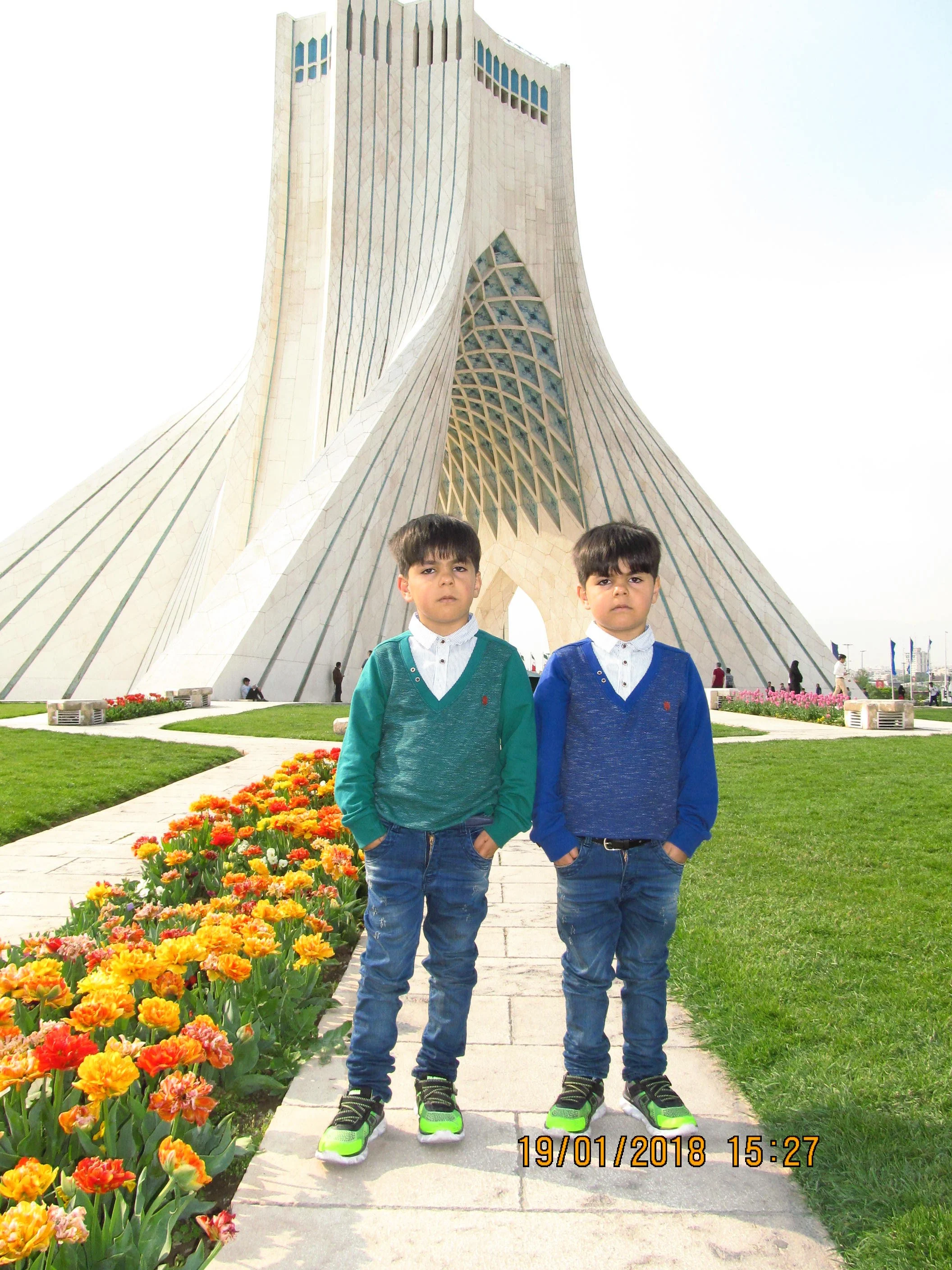 Two boys standing on a walkway with colorful flowers in front of the Azadi Tower in Iran.