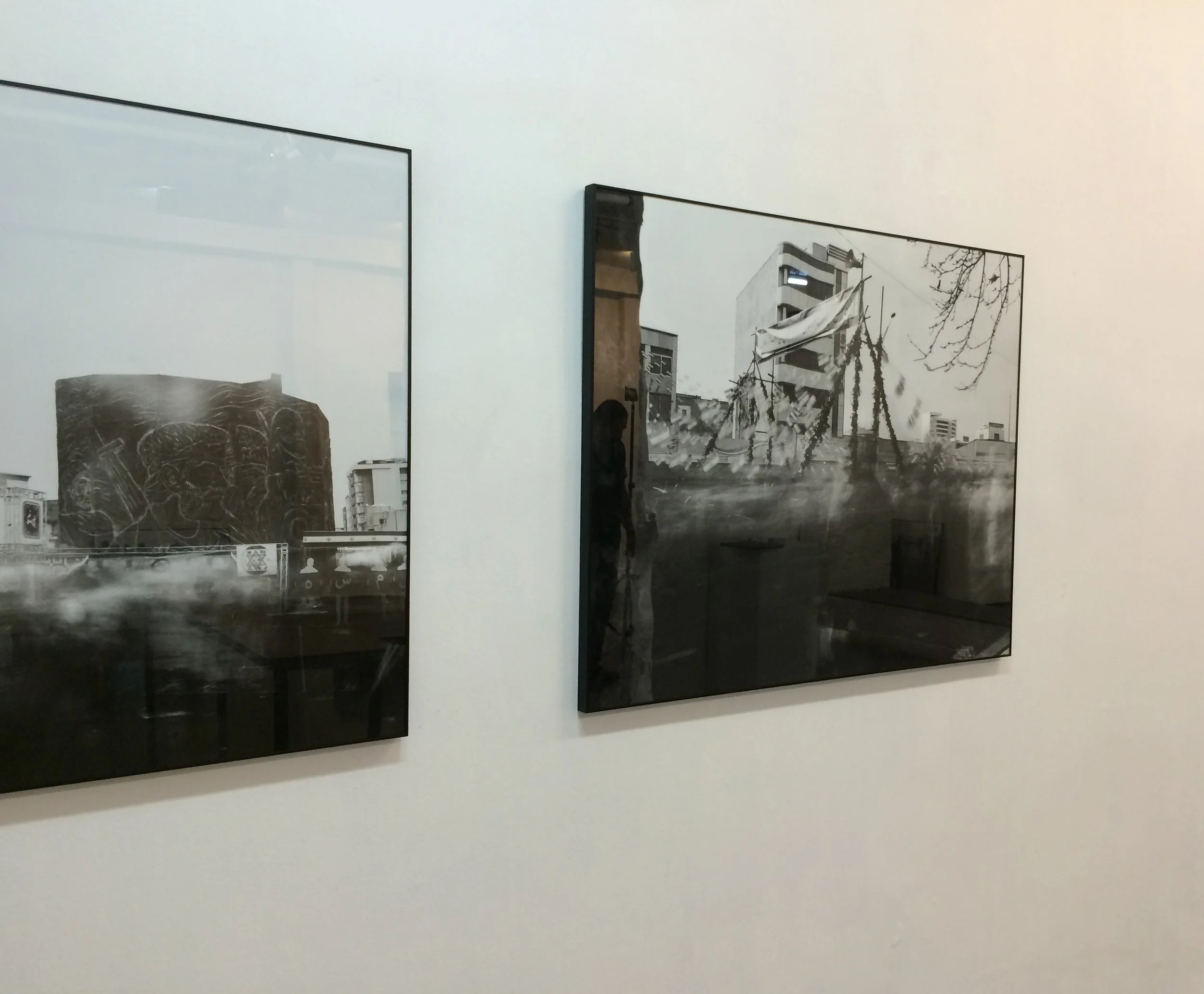 Black and white photographs displayed on a white gallery wall, reflecting indoor and outdoor scenes, including buildings, trees, and urban landscapes.