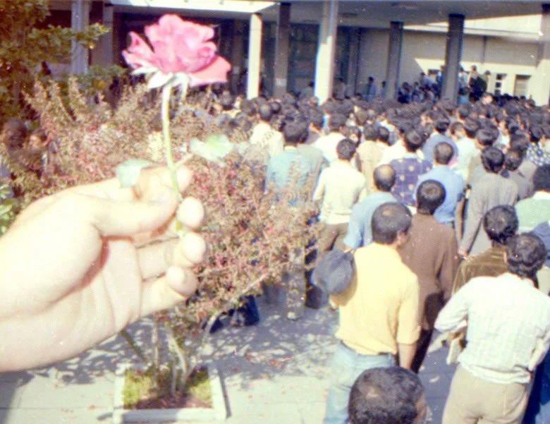 A hand holding a small bouquet with a pink flower, overlooking a crowd of people gathered outside a building.