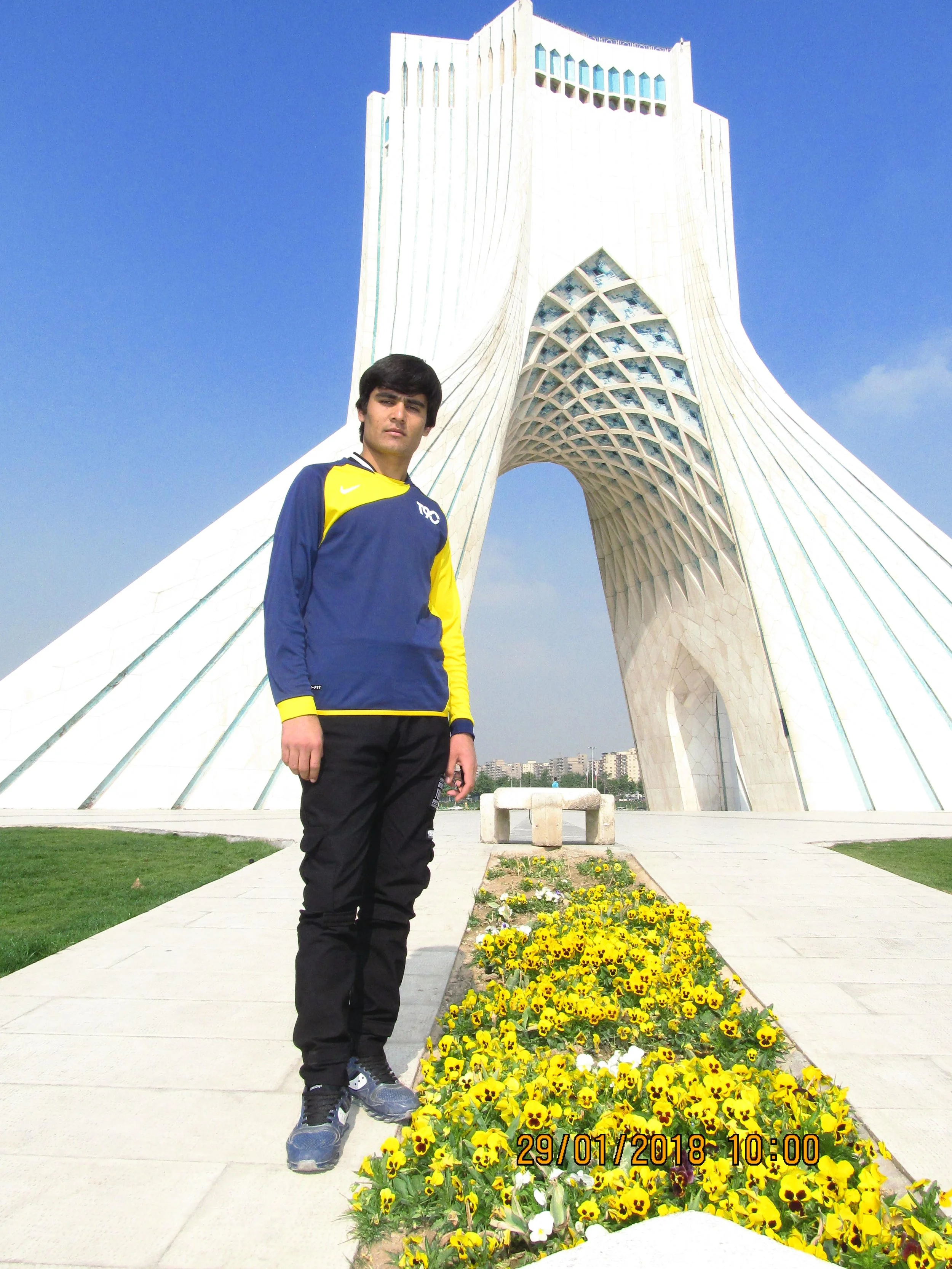 A young man standing in front of the Azadi Tower in Tehran, Iran, with a pathway of yellow flowers leading towards the monument on a sunny day.