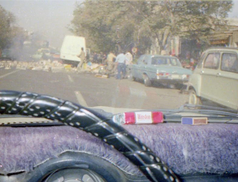 View from inside a car looking at a street scene with old cars, a white van, and people walking near a sidewalk, with a tree-lined background.
