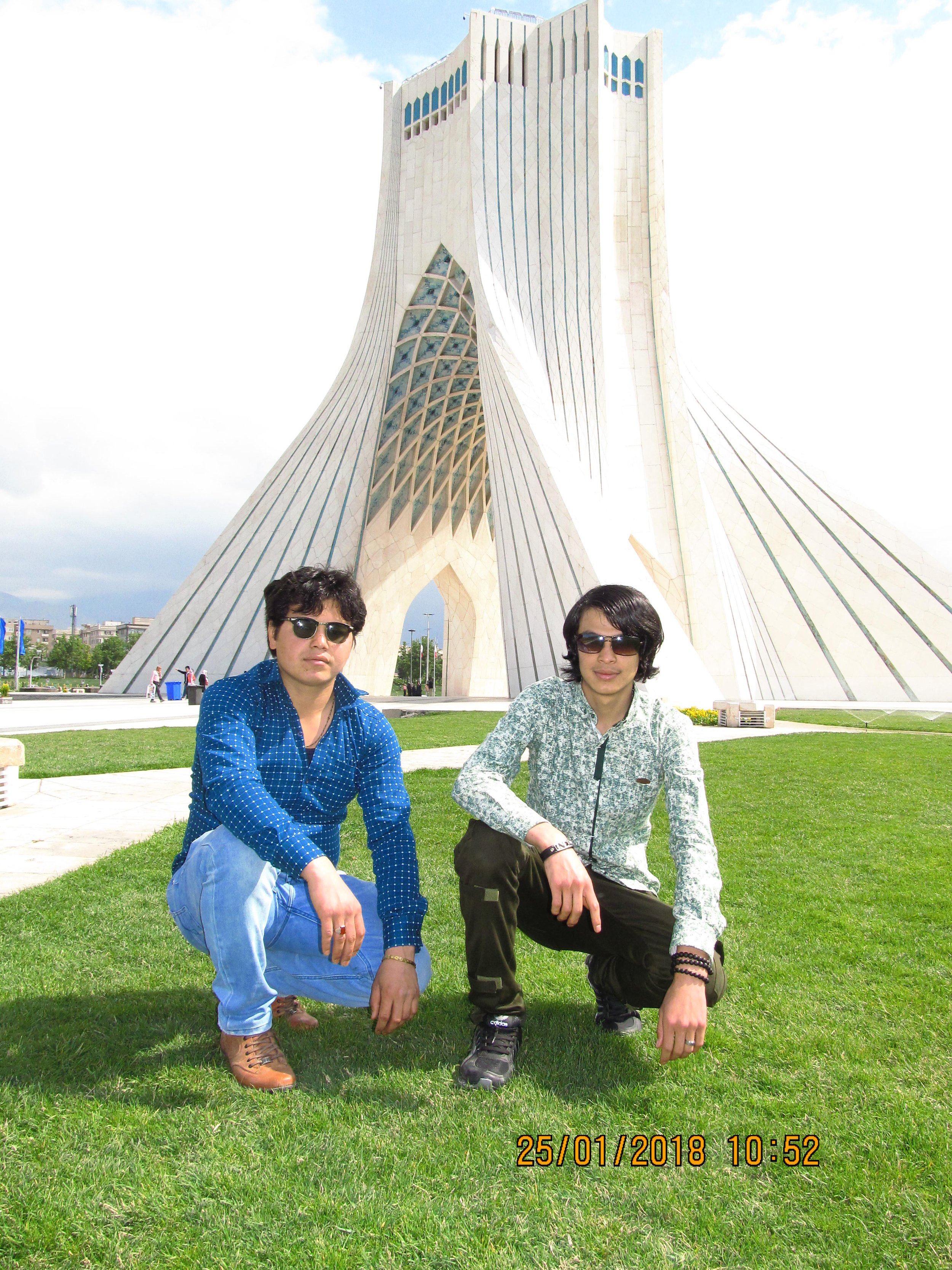 Two young men squatting on grass in front of a modern architectural building, wearing sunglasses and casual clothing, with a clear sky above.