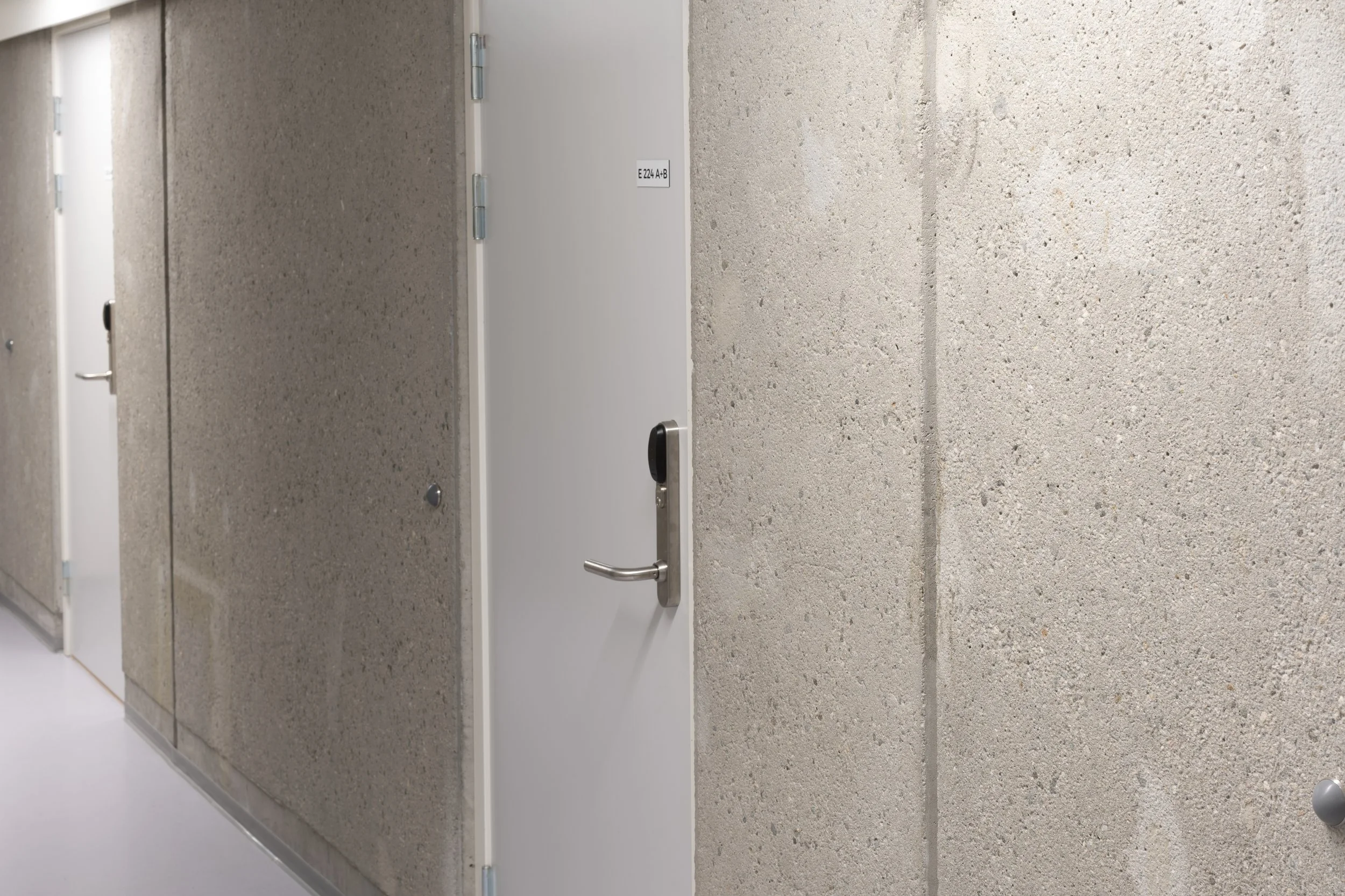 A corridor with concrete walls, featuring a closed white door with a digital lock and a small sign that reads 'E24 A-B'.