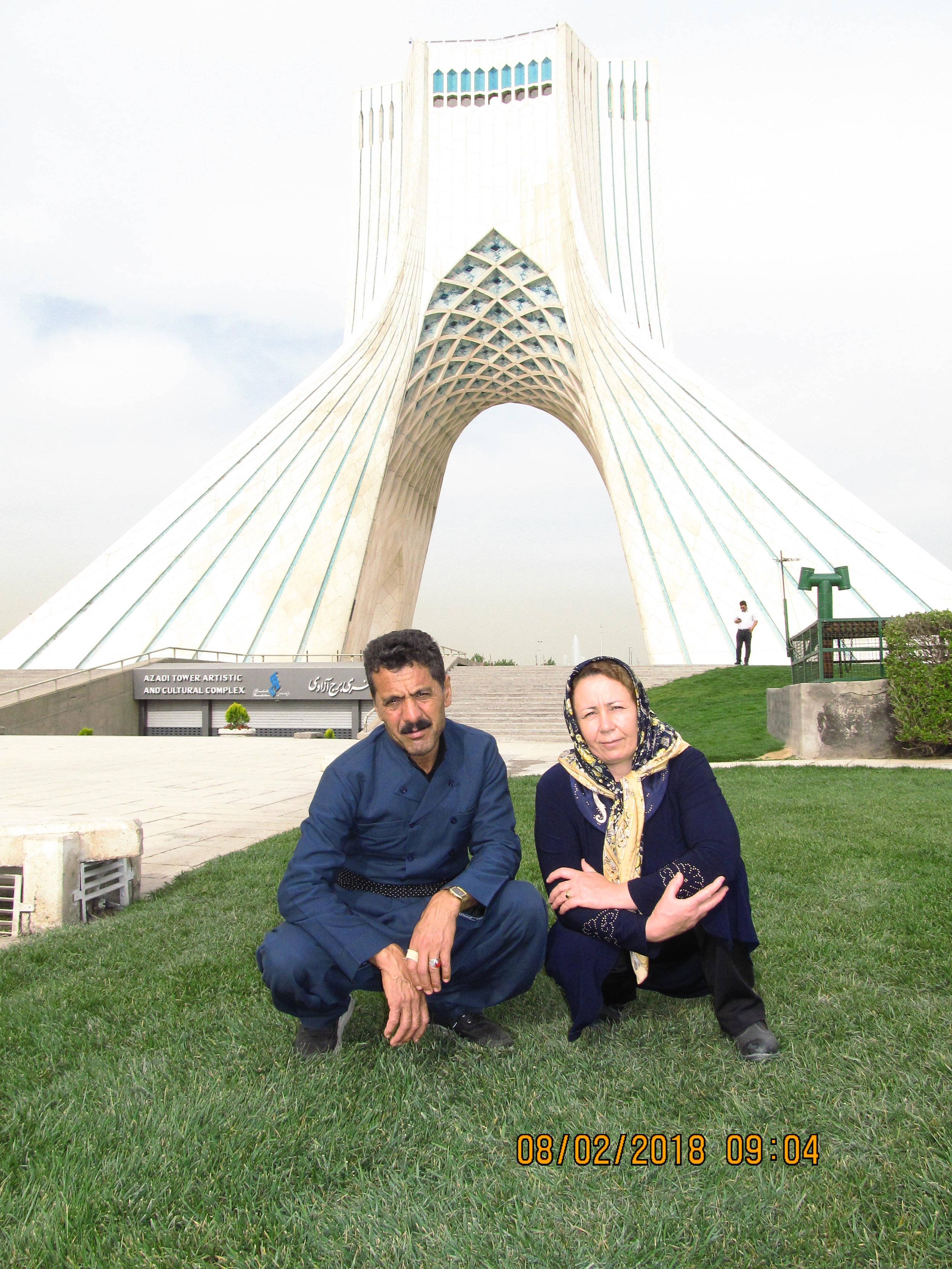 A man and woman crouching on grass in front of Azadi Tower in Tehran, Iran, with a cloudy sky above.