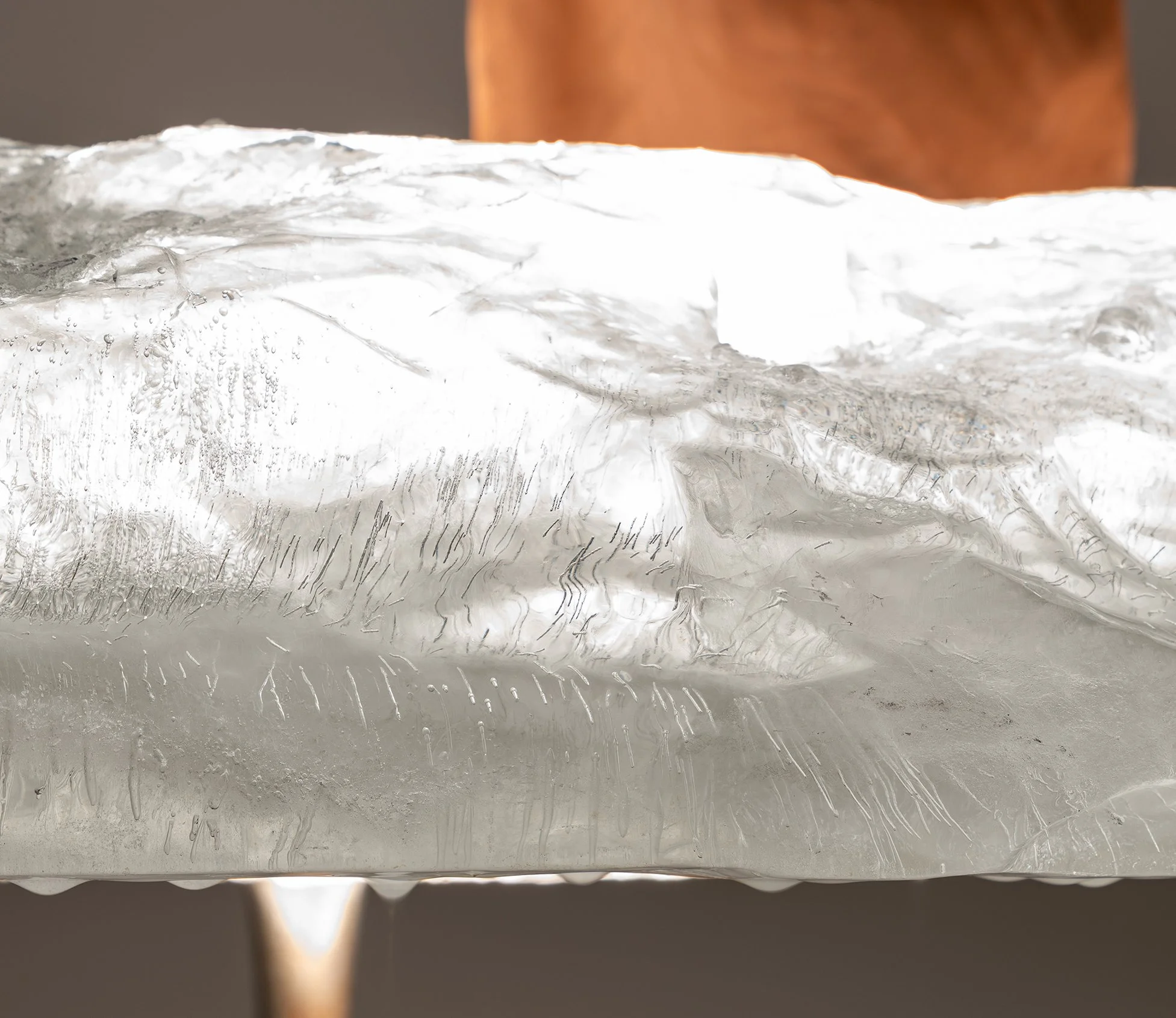 Close-up of a transparent ice block with water droplets and textured surface, with a blurred wooden object in the background.