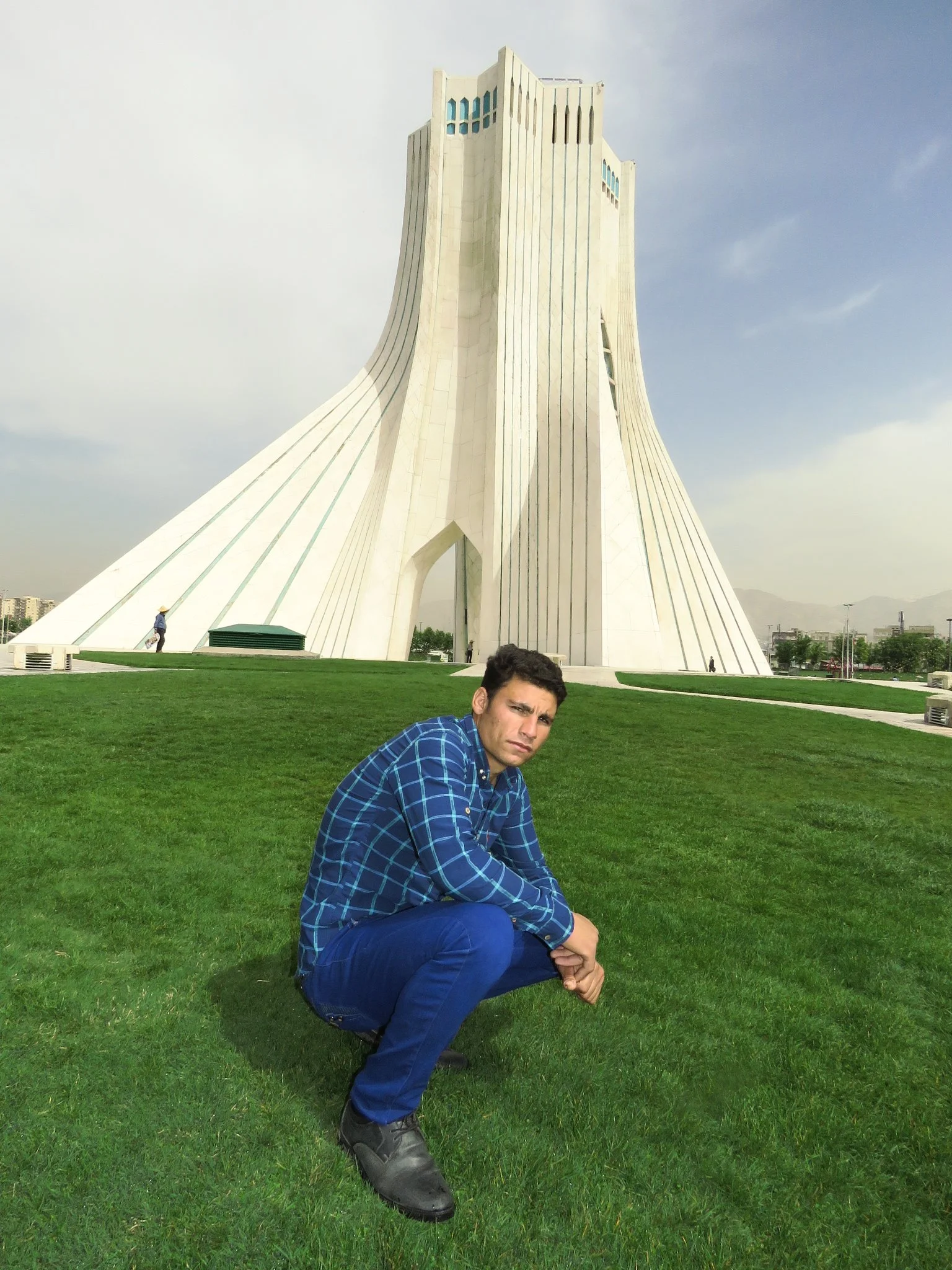 A man in a blue plaid shirt crouching on green grass in front of the Azadi Tower in Tehran, Iran.