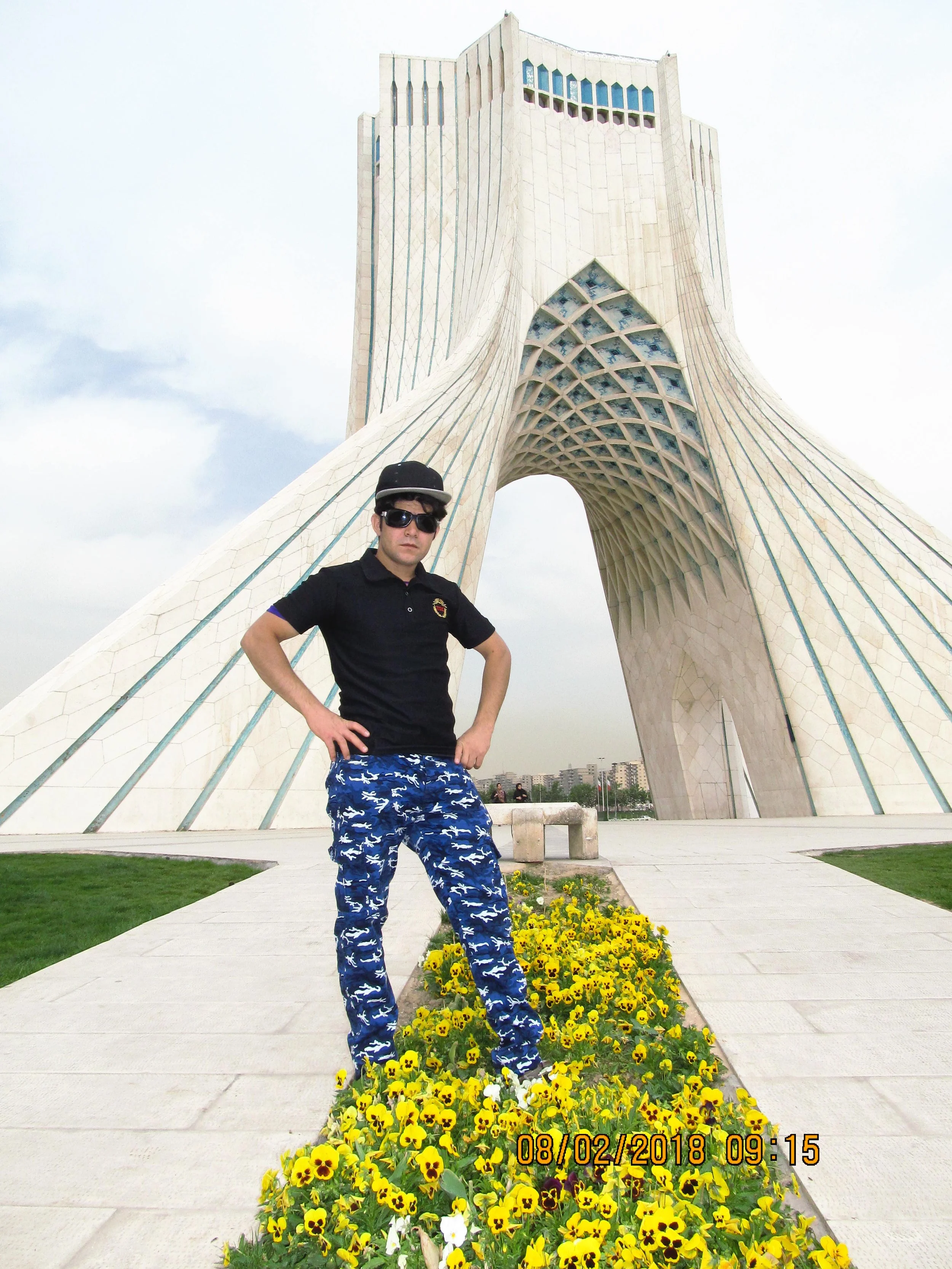 A young man standing in front of Azadi Tower in Tehran, Iran, wearing a black polo shirt, blue patterned pants, sunglasses, and a cap, with hands on hips, on a sunny day with partly cloudy sky, beside a flower bed of yellow and purple flowers.