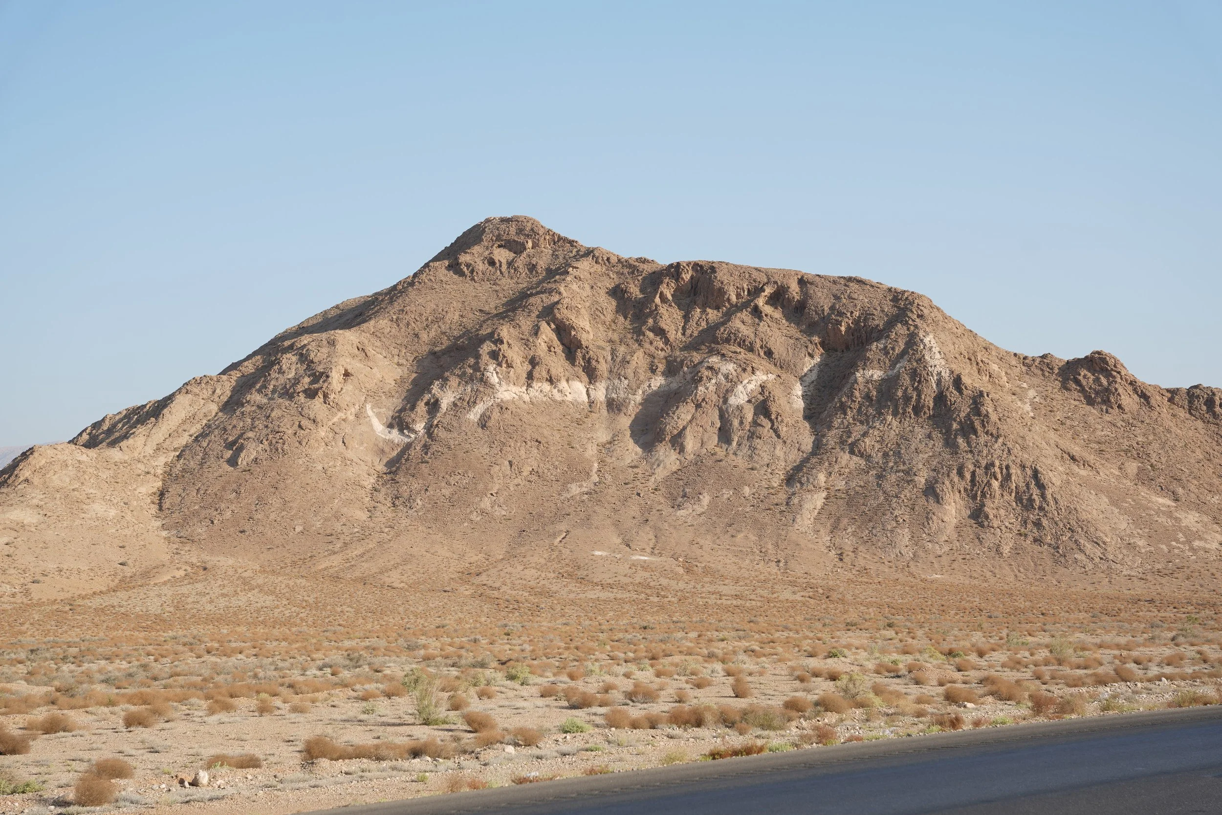 A rugged desert mountain with sparse vegetation in the foreground and a clear blue sky.