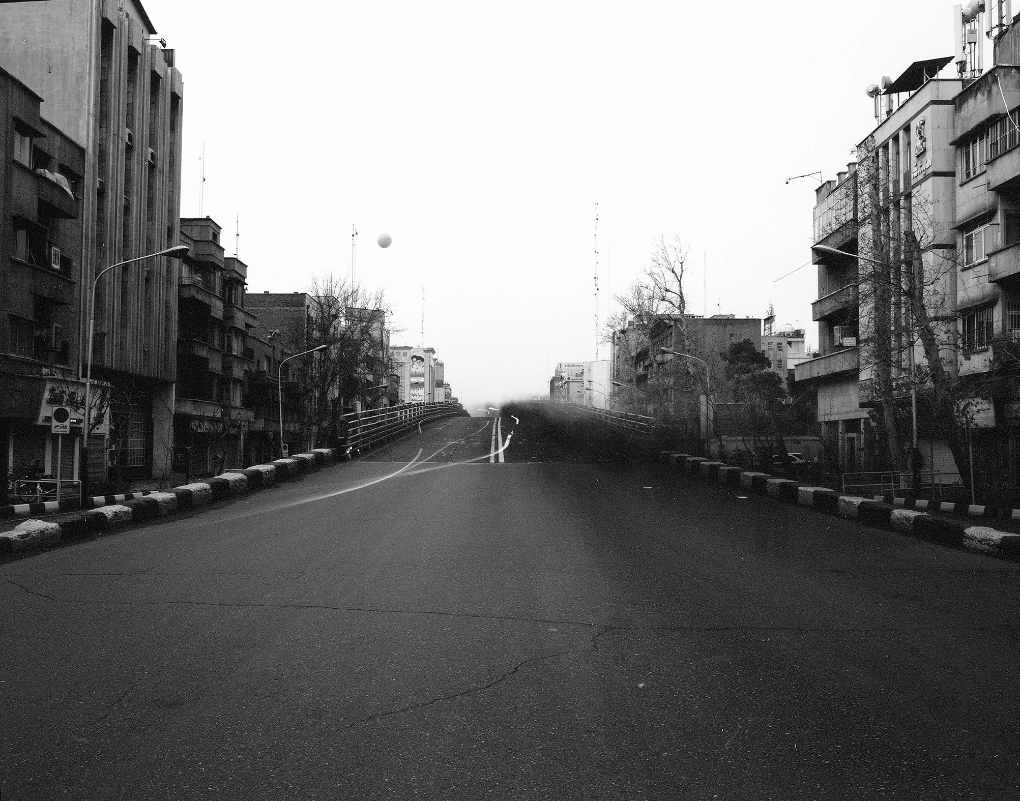 Empty city street with buildings on both sides, leafless trees, and a single floating ball in the sky, black and white photo.