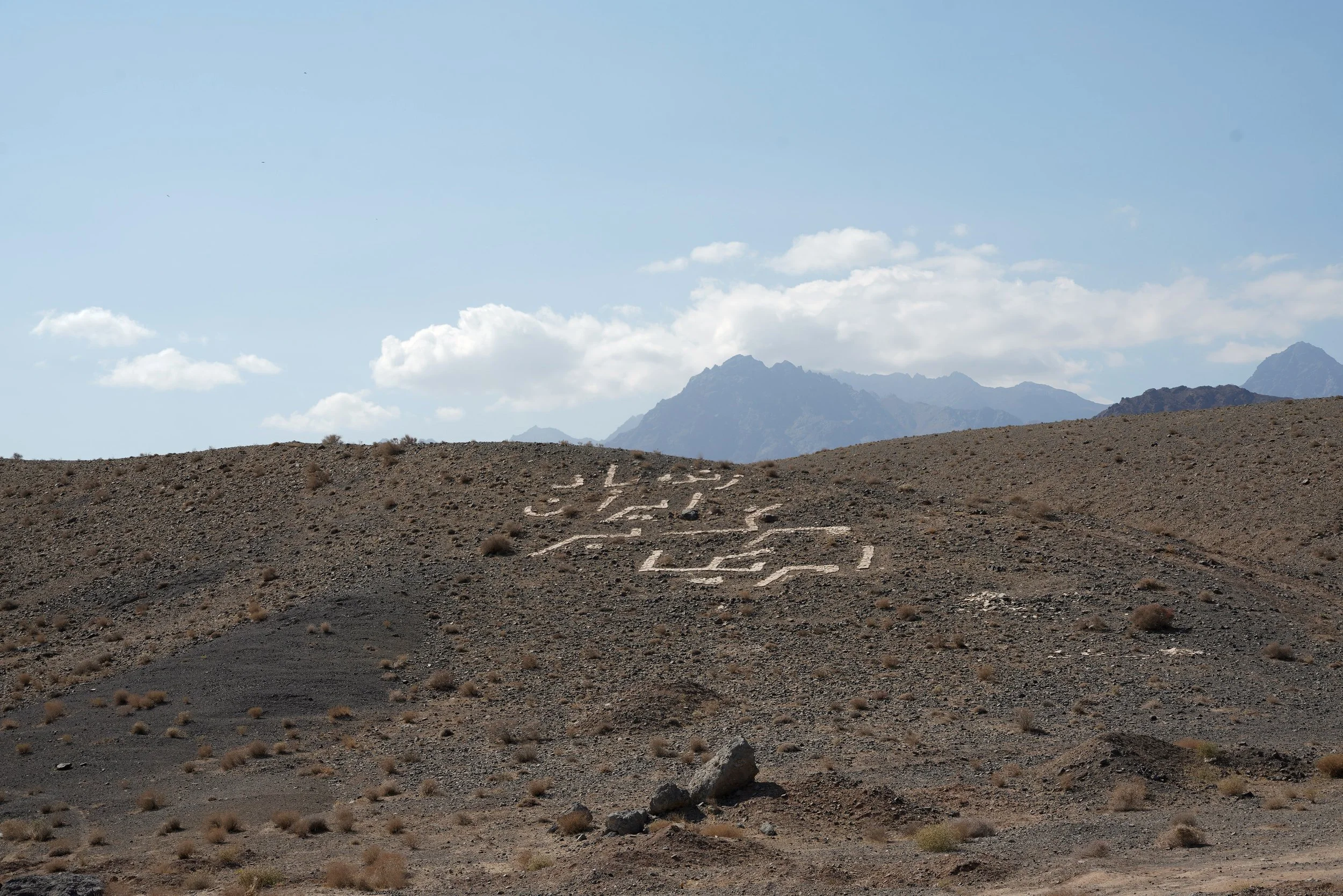 Remote desert landscape with mountains in the background and a large white geometric symbol or pattern on the ground in the foreground.