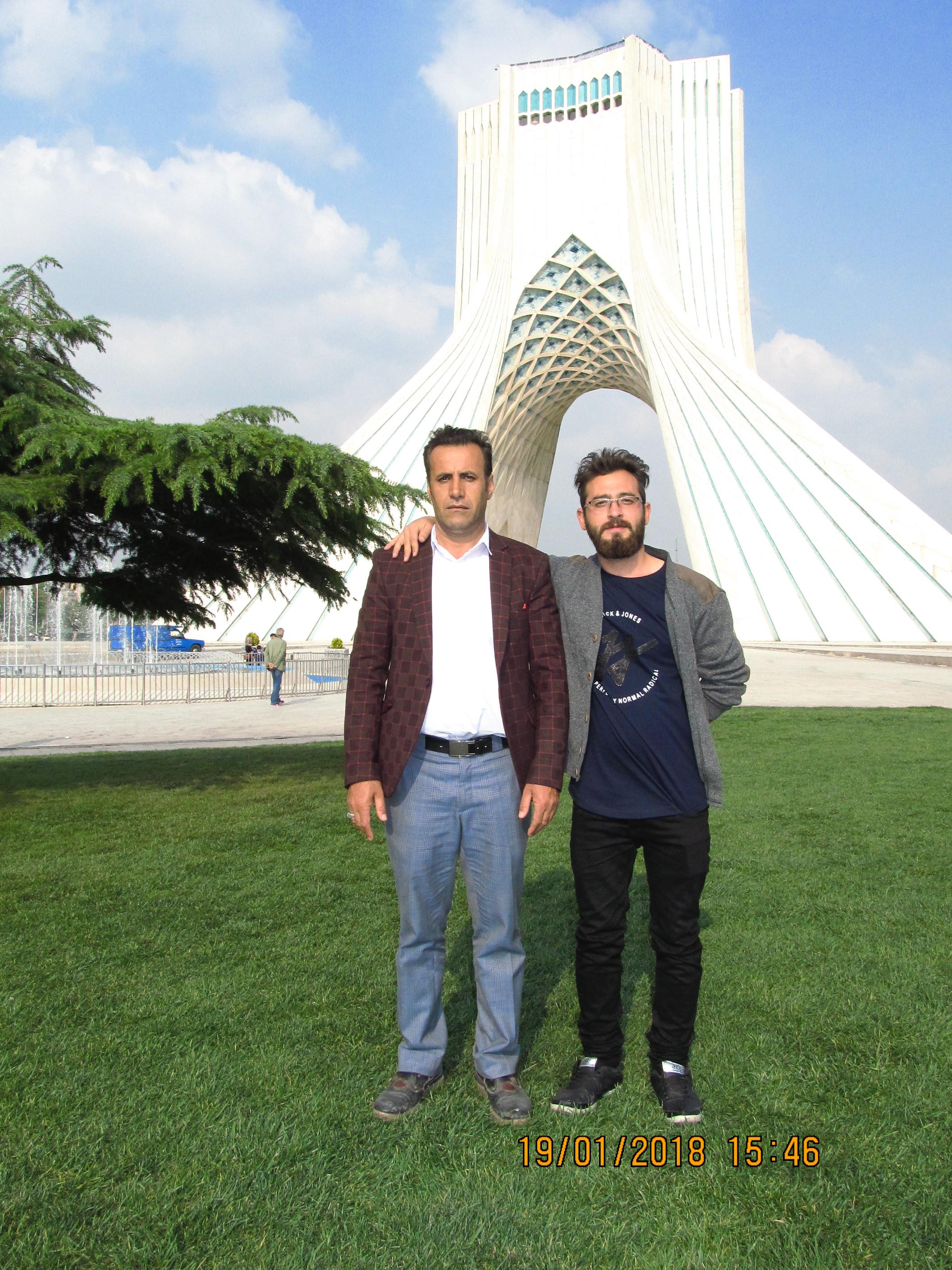 Two men standing in front of Azadi Tower in Tehran, Iran, with green grass and a tree on the left side, and a clear sky with some clouds in the background.