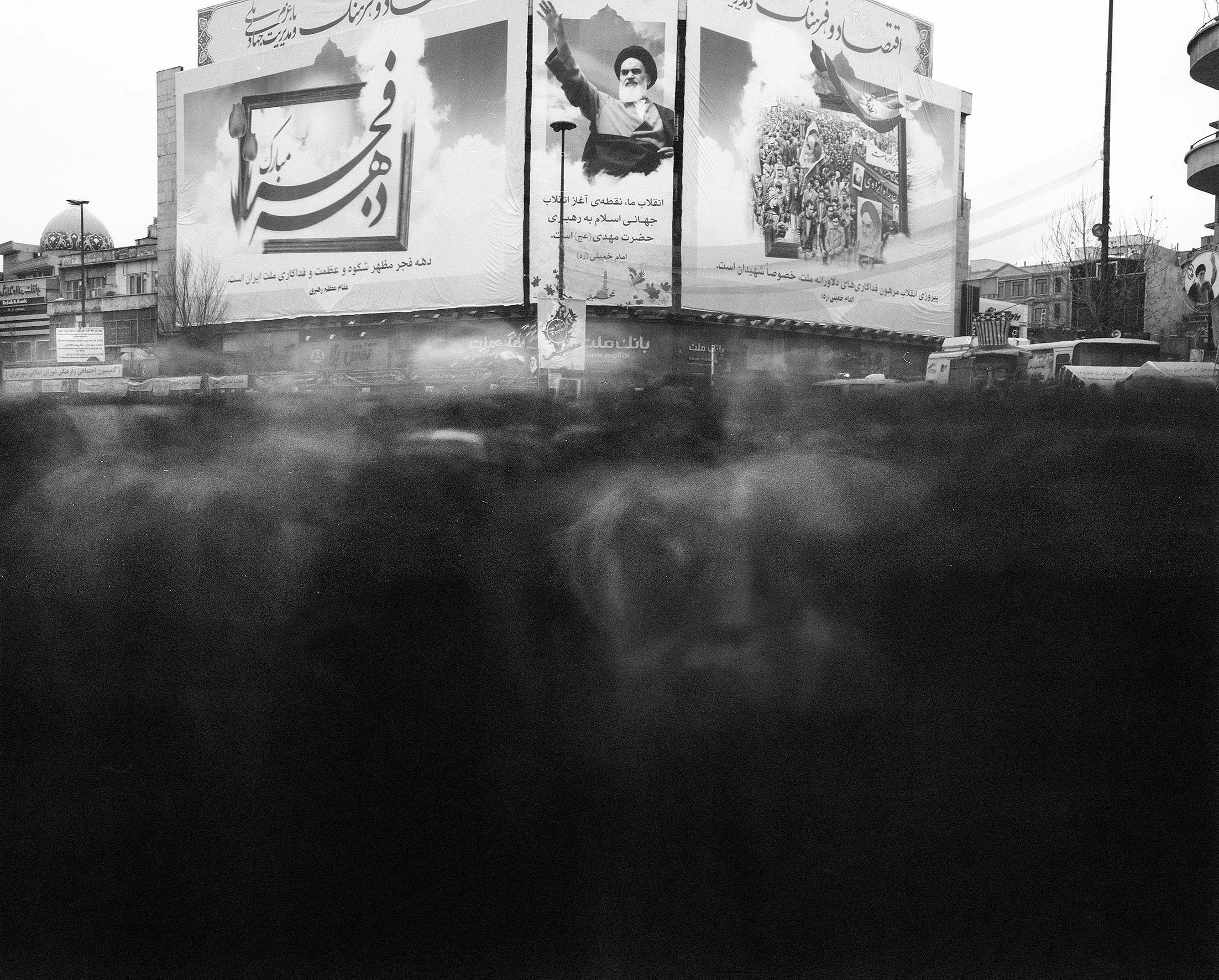 Black and white photo of large billboards with Persian text and images, including a man with a raised hand. The billboards are on a city street with buildings, trees, and cars in the background.
