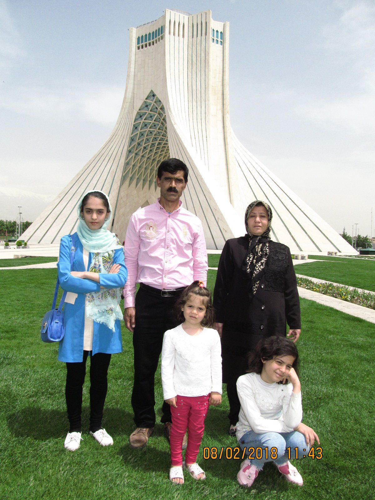 A group of six people, including two children, posing outside in front of the Azadi Tower in Tehran, Iran. The group includes a man, two women, and three girls, all standing and sitting on a well-manicured lawn.