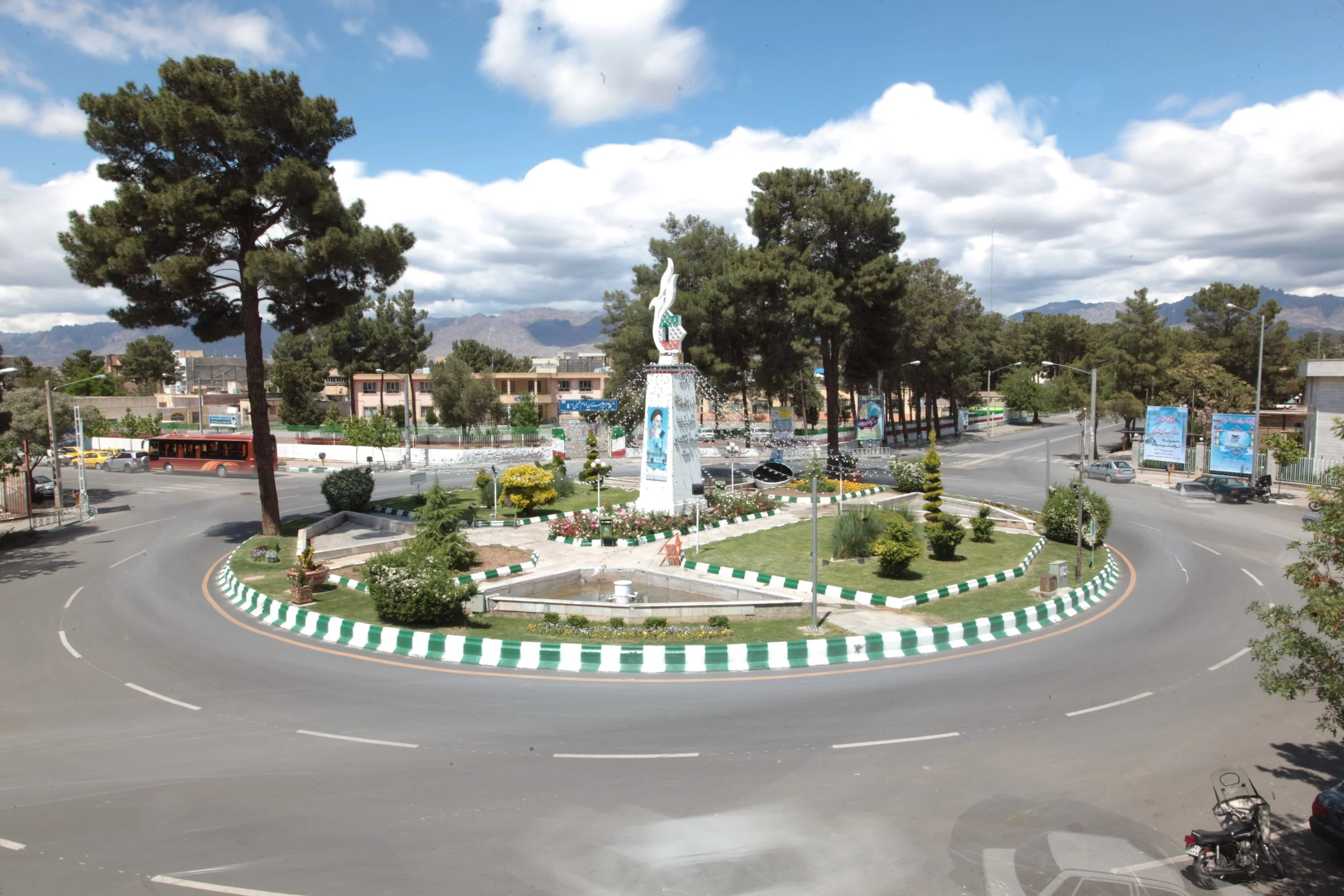 A roundabout with a central monument and landscaped gardens, surrounded by a gray road, trees, and buildings in the background, with mountains and partly cloudy sky in the distance.
