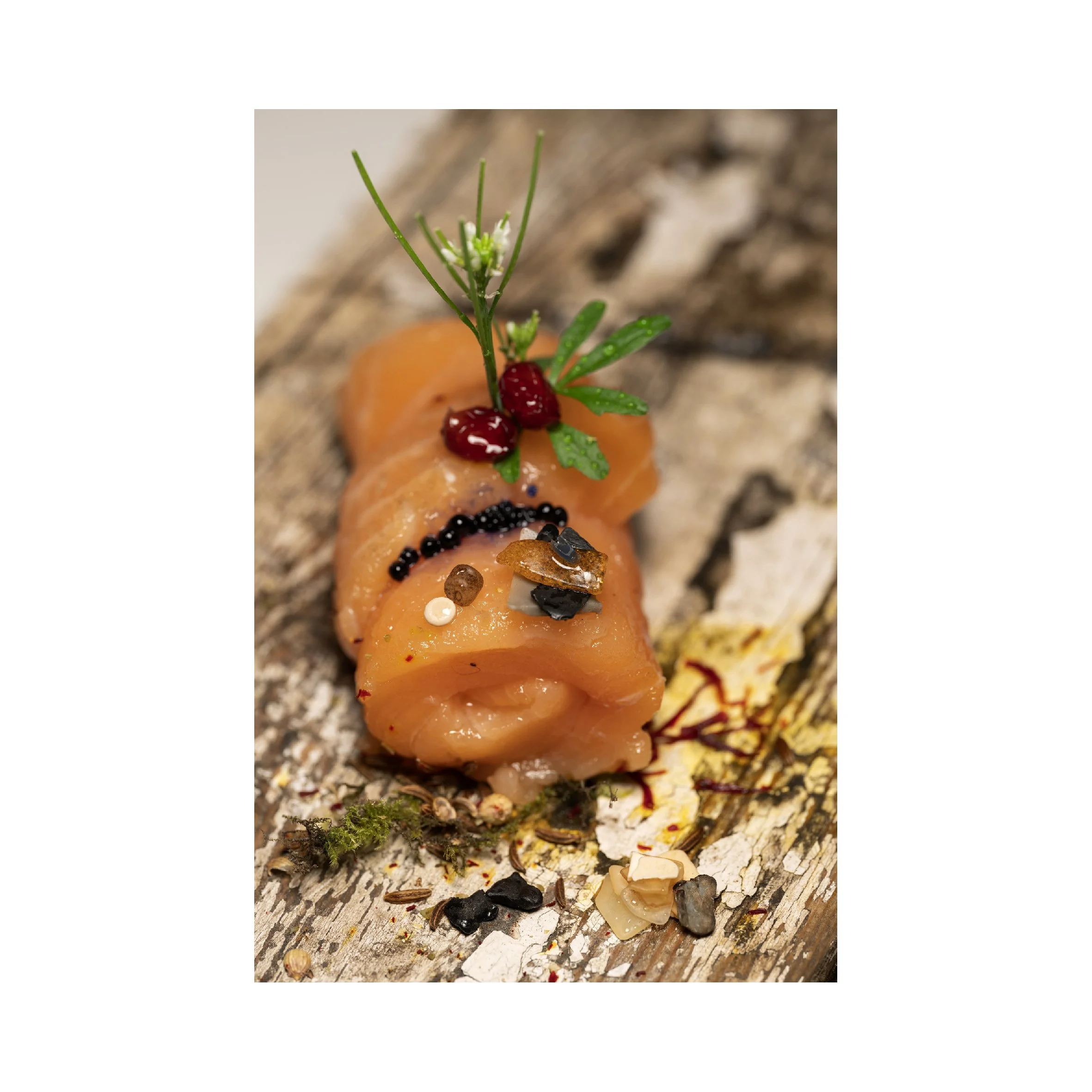 Close-up of a dish with rolled slices of salmon garnished with herbs, berries, and spices on a rustic wooden surface.