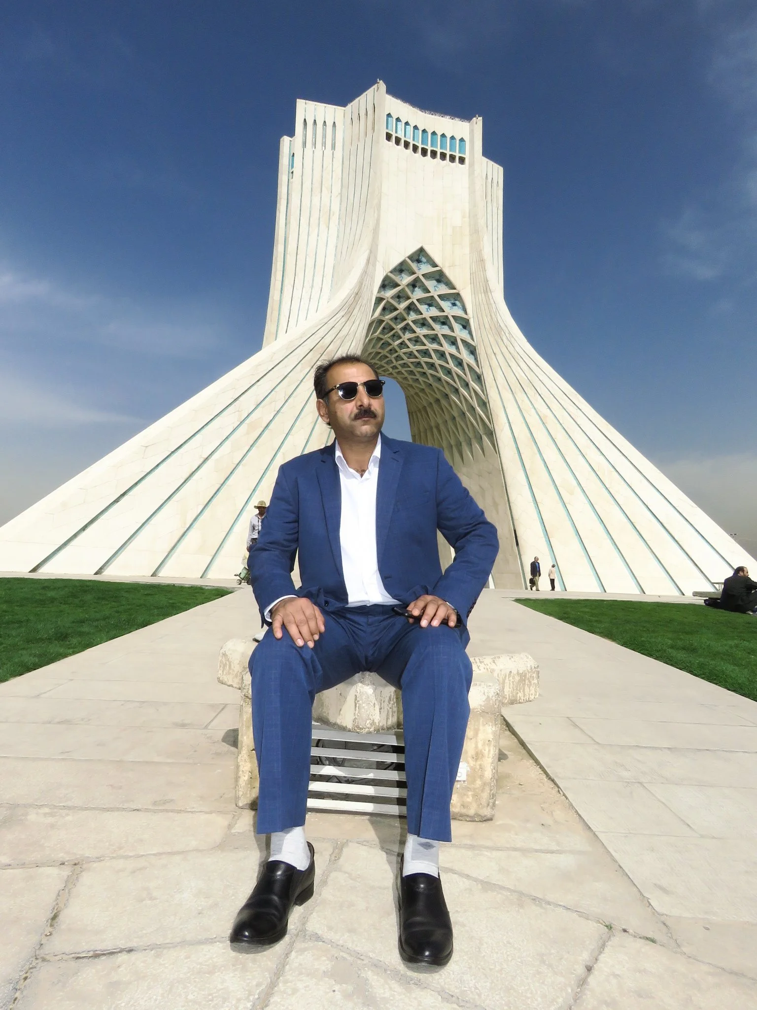 A man in a blue suit, white shirt, black shoes, and sunglasses sitting on a stone bench in front of the Azadi Tower in Tehran, Iran, with a clear blue sky in the background.