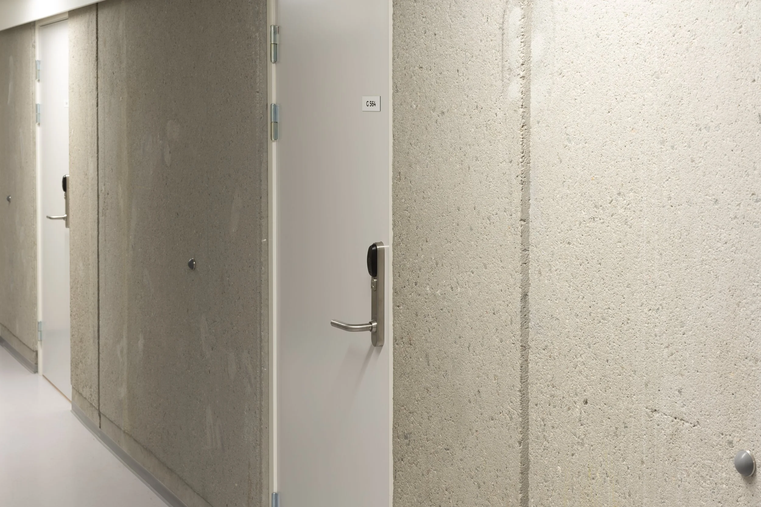 Hallway with concrete walls and multiple closed white doors, one with a black handle.